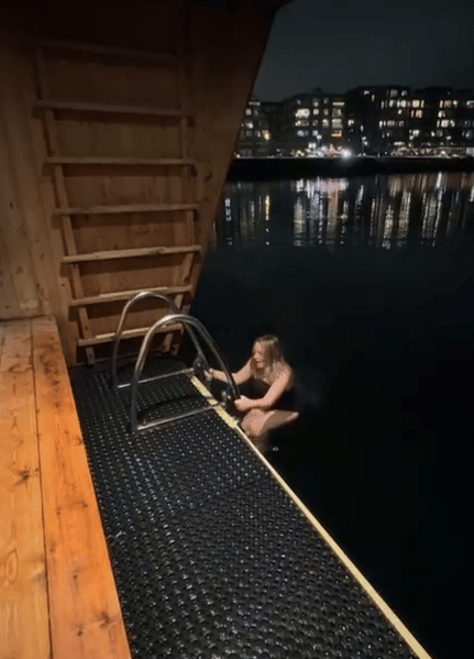 Someone sits on a floating dock at night with city lights reflecting on the dark water in Stockholm.