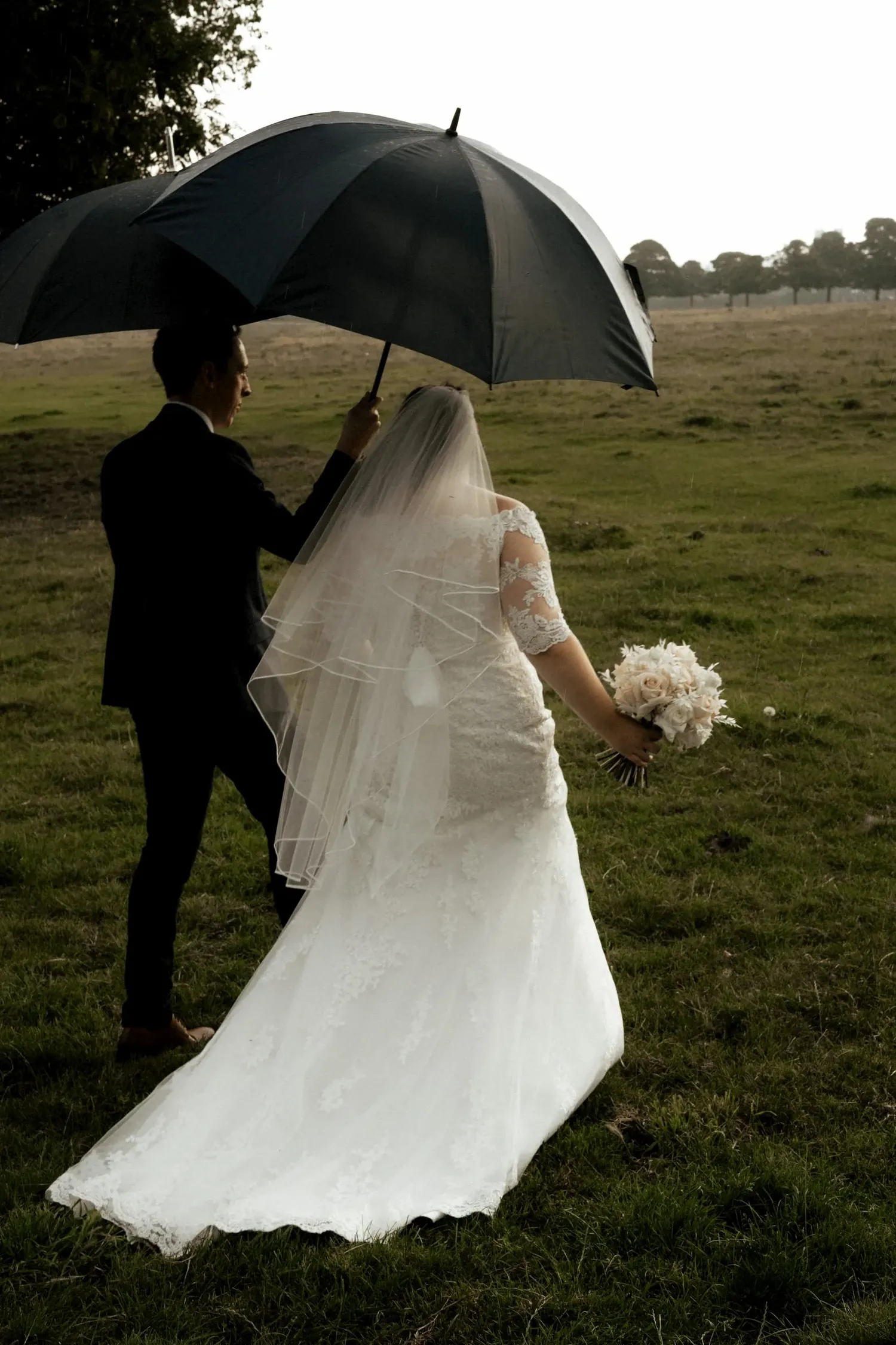 Couple shares intimate moment under black umbrella in countryside wedding photo.
