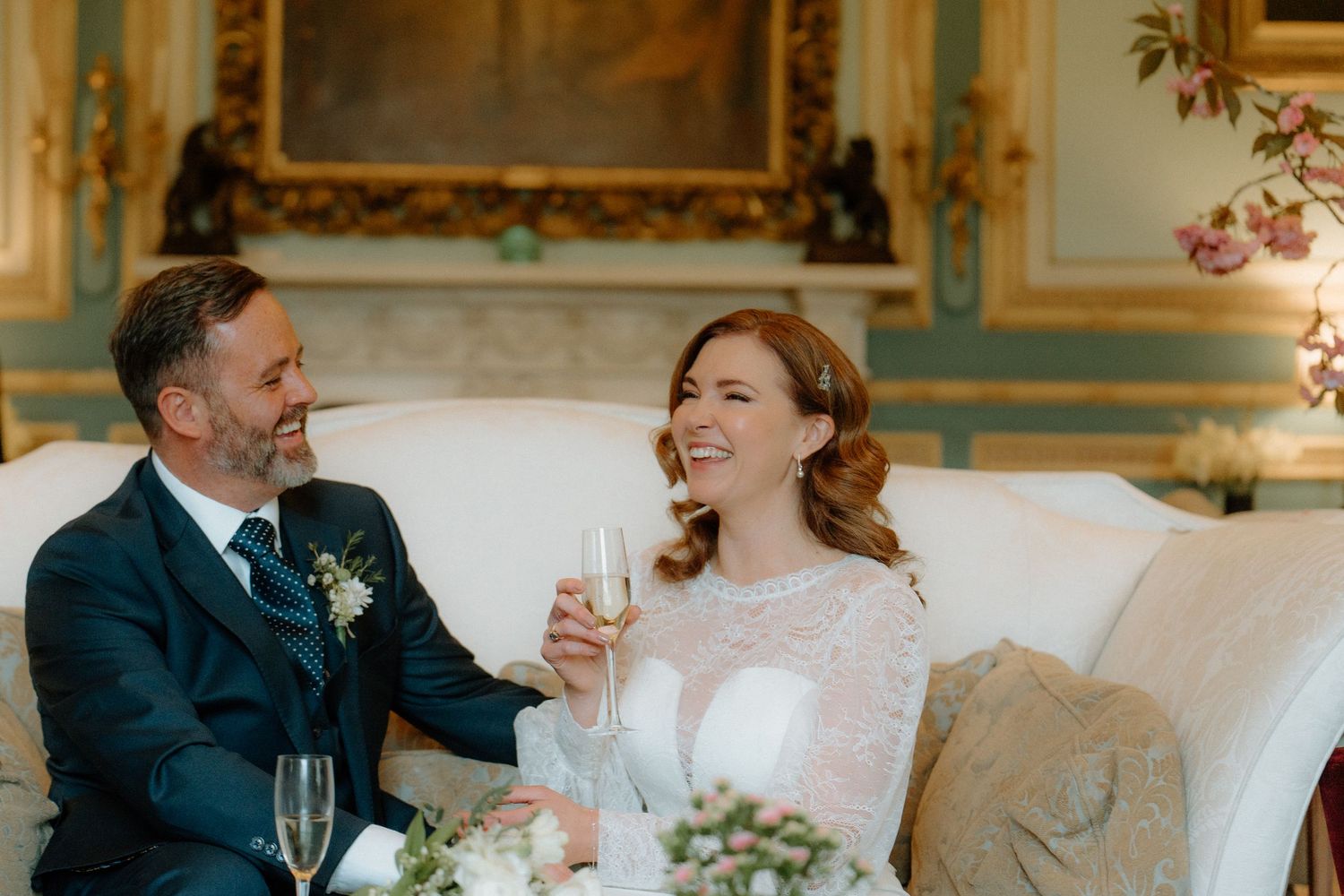 Bride and groom laughing and toasting with champagne on elegant sofa in ornate wedding venue.
