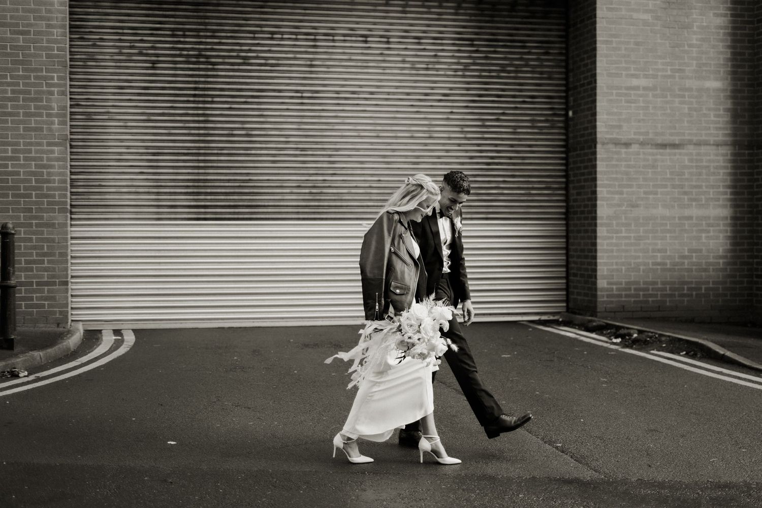 Black and white photo of a bride and groom walking past a brick wall and roller door on their wedding day.