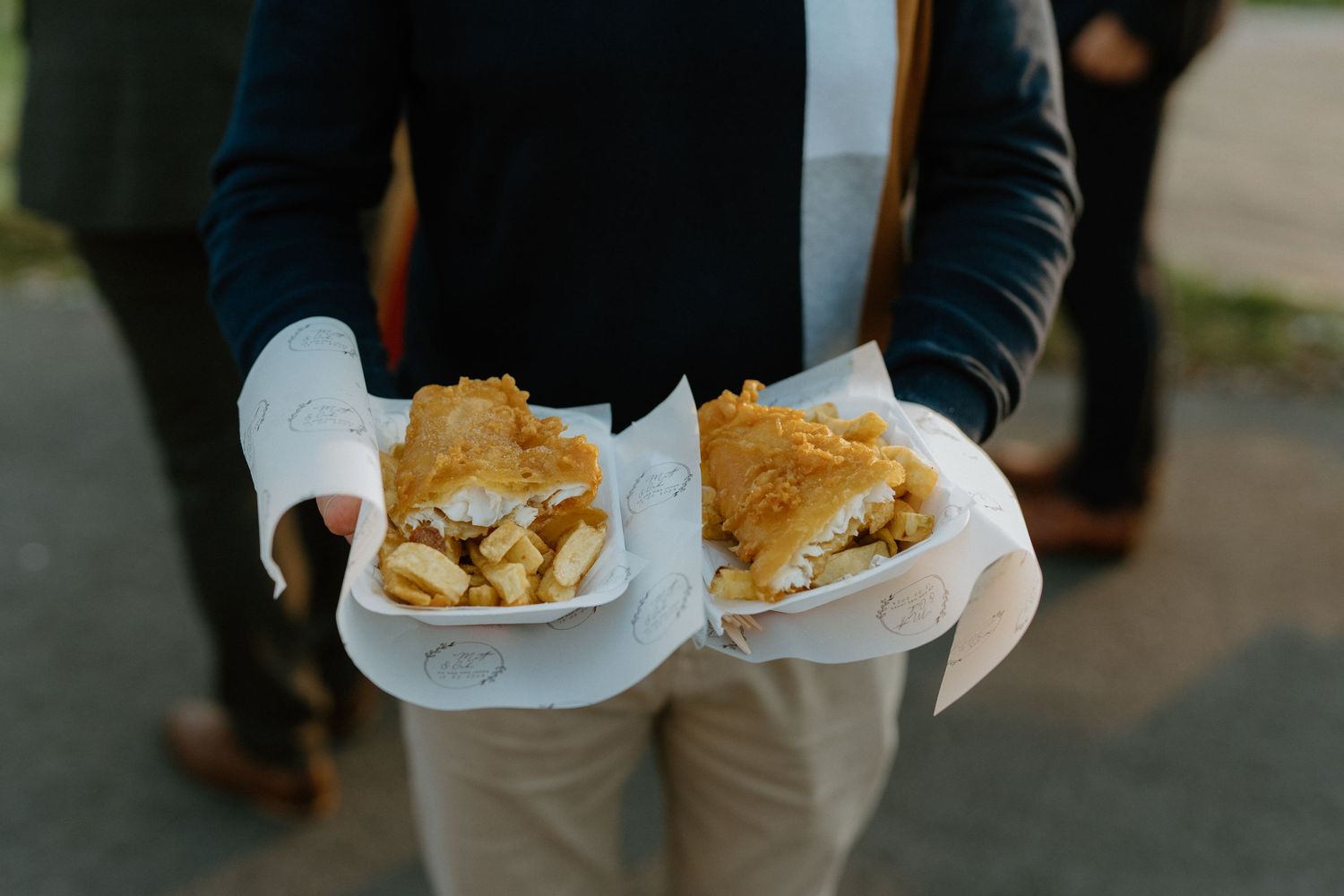 Hands wearing white gloves holding two paper trays of fries topped with crispy breadcrumbs or fried onions.