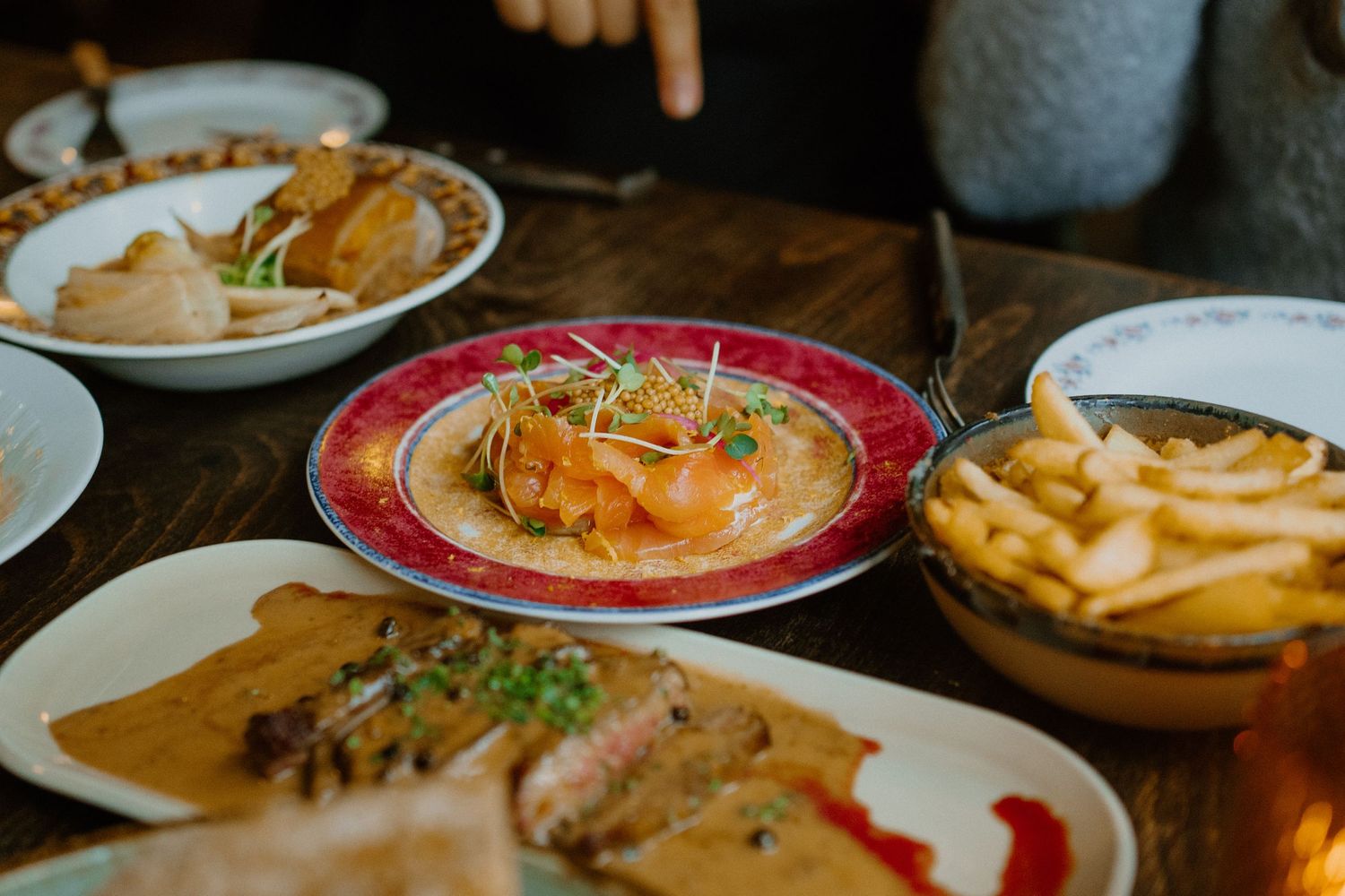 Multiple plates of restaurant food including french fries and meat dishes served on a wooden table in warm lighting.