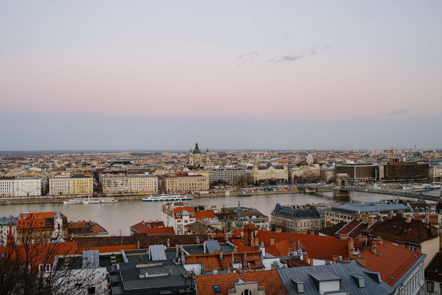 Panoramic cityscape of Budapest at sunset showing the Danube River and iconic red rooftops against a pastel sky.