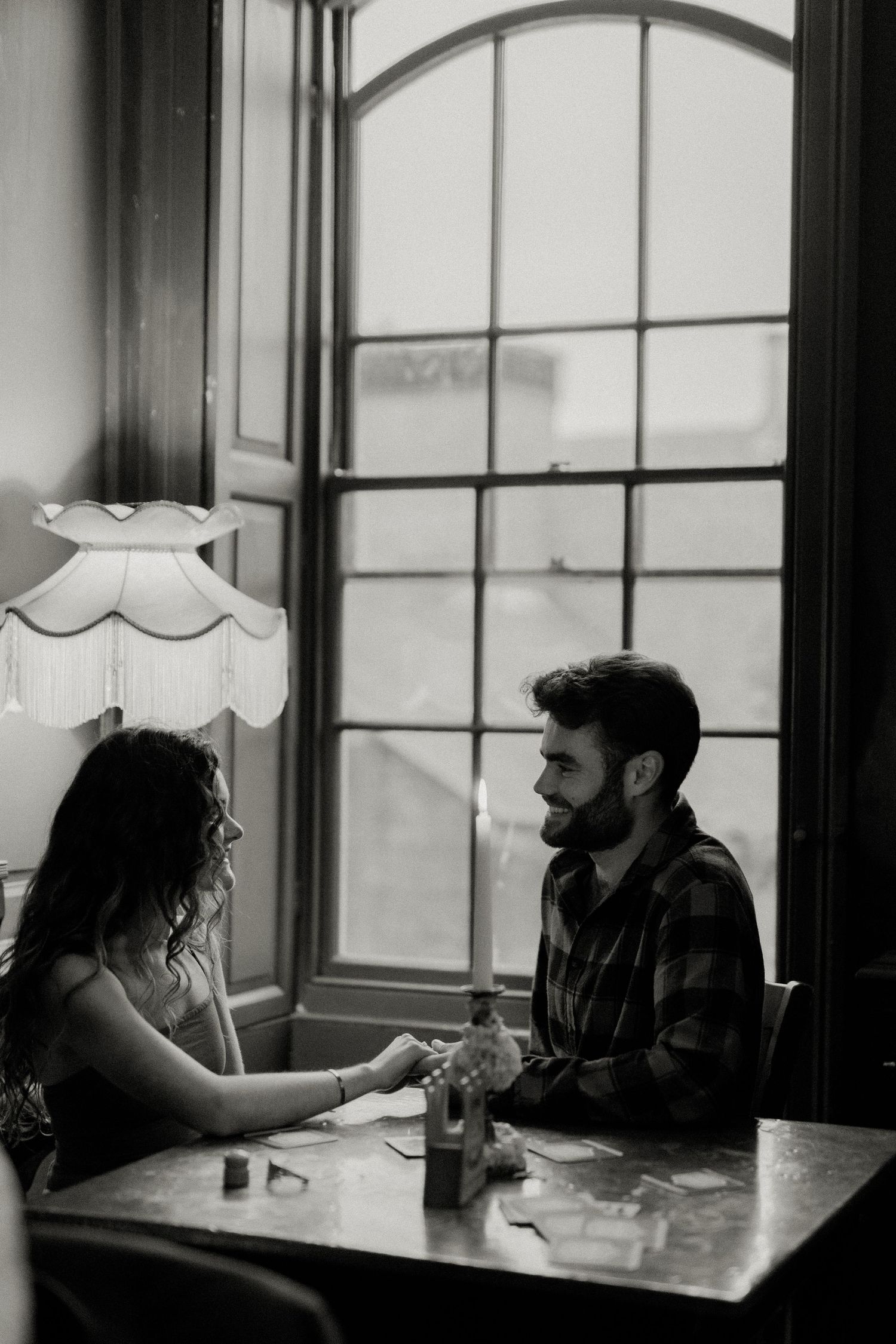 A couple sits together at a dimly lit table by an arched window, sharing an intimate conversation in black and white.