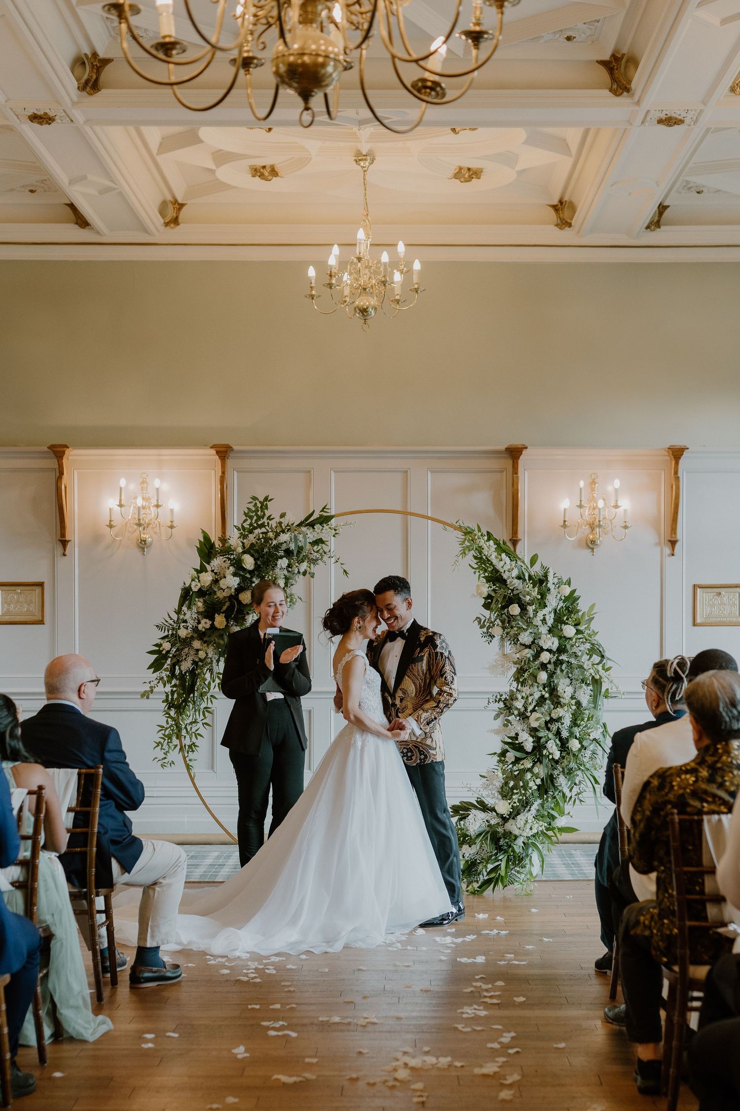 Bride and groom share first kiss under floral arch in elegant ballroom wedding ceremony.