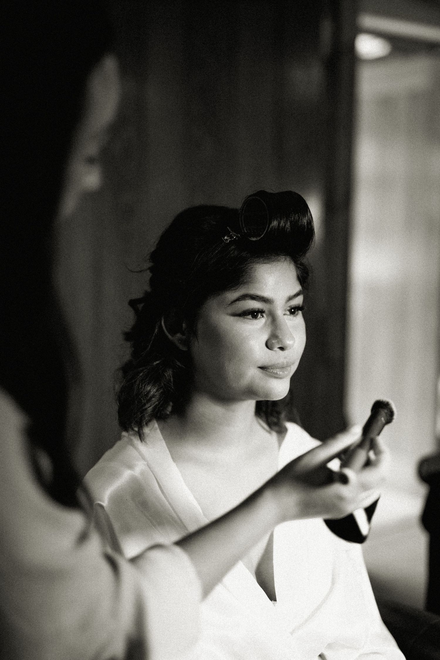 A black and white portrait of a bride having her makeup applied while looking in a mirror before her wedding.
