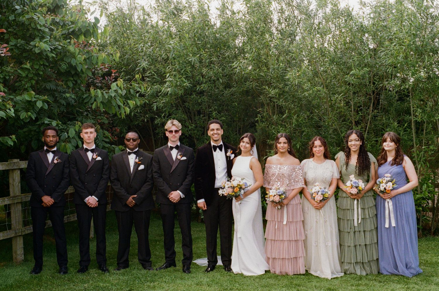 Wedding party poses outdoors with groomsmen in black suits and bridesmaids in mixed pastel gowns by greenery.