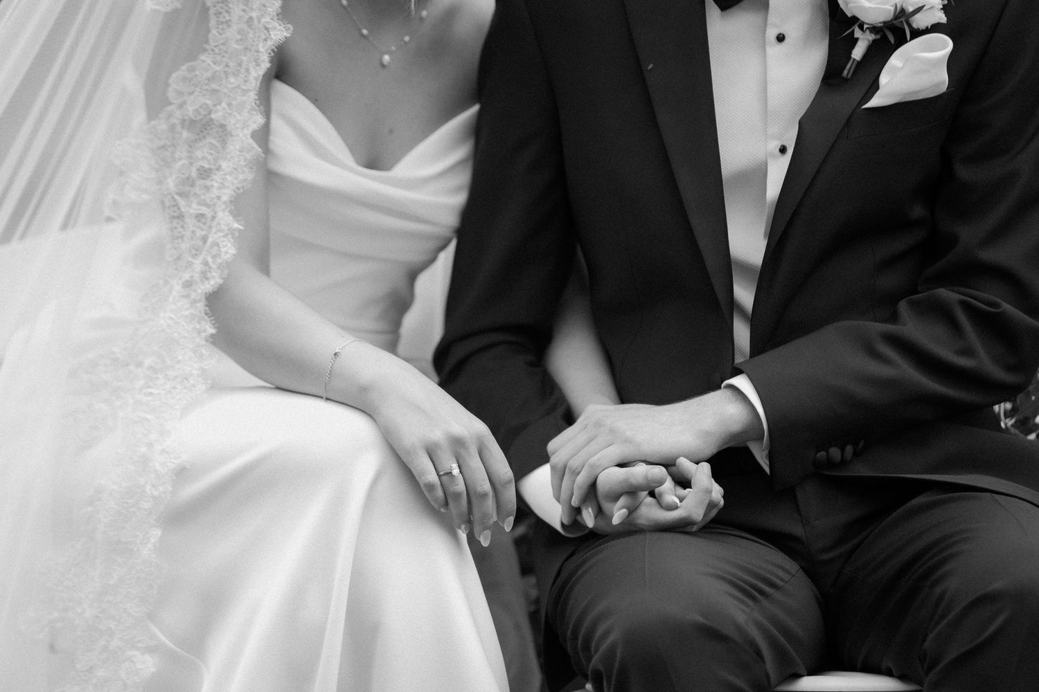 Black and white close-up of bride and groom holding hands while seated during their wedding ceremony.