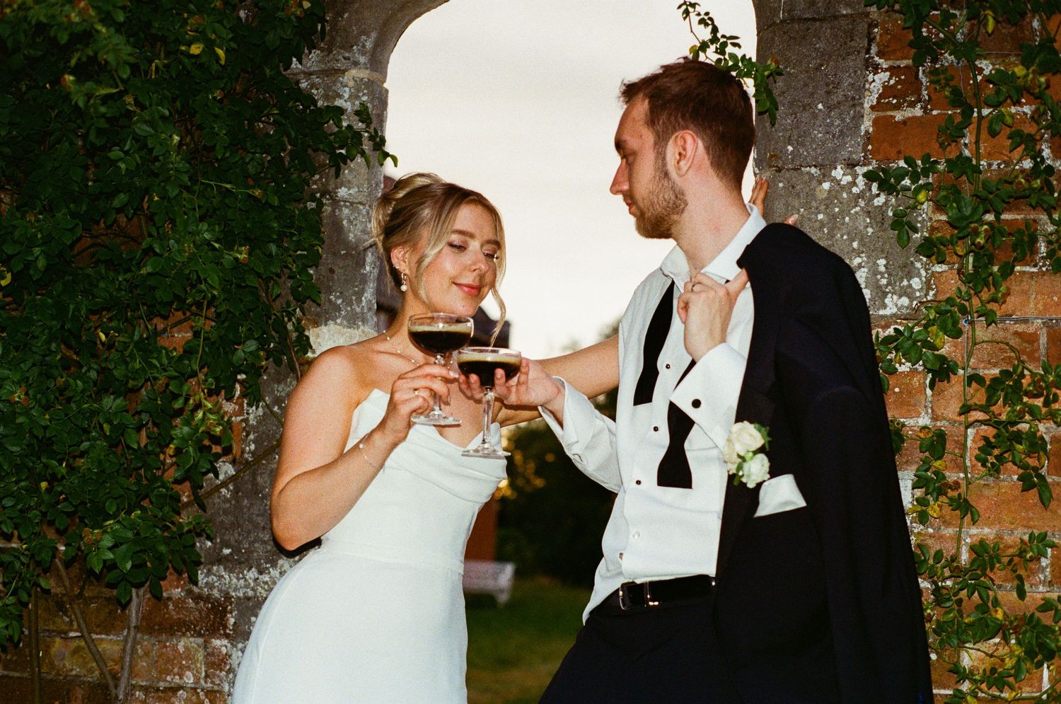 Smiling bride and groom toasting with red wine glasses at rustic stone arch wedding venue.