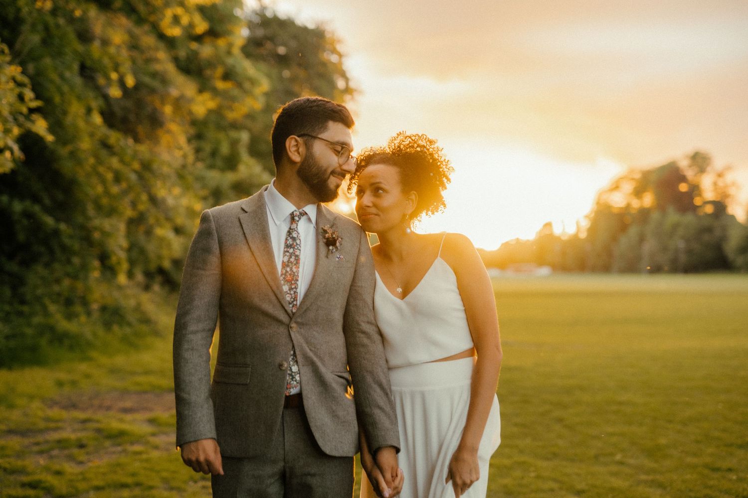 Couple sharing a romantic moment at golden sunset in a green meadow, bride in white dress, groom in grey suit.