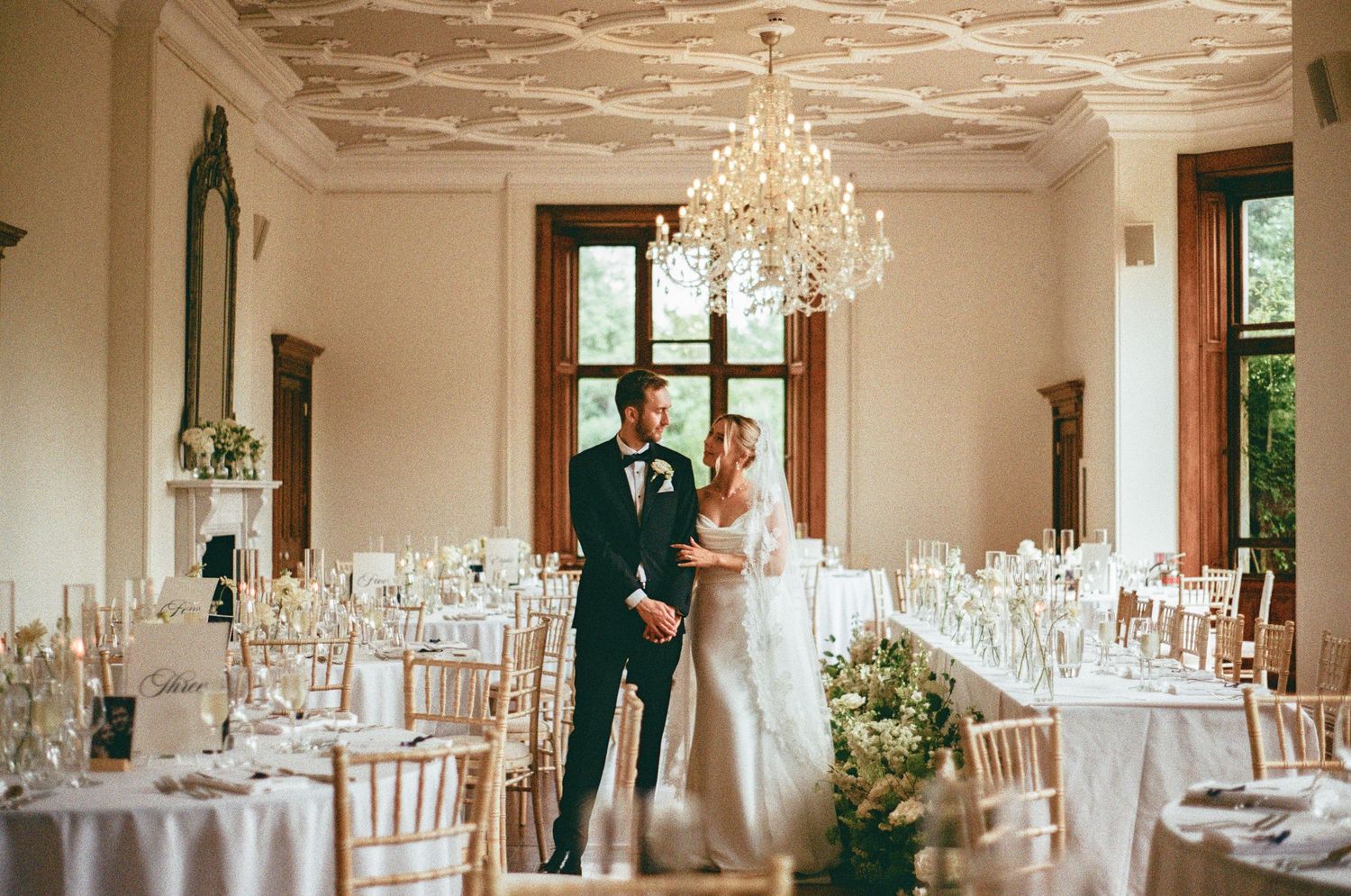 Bride and groom share romantic moment in elegant chandelier-lit reception hall with gold chairs and white tables.