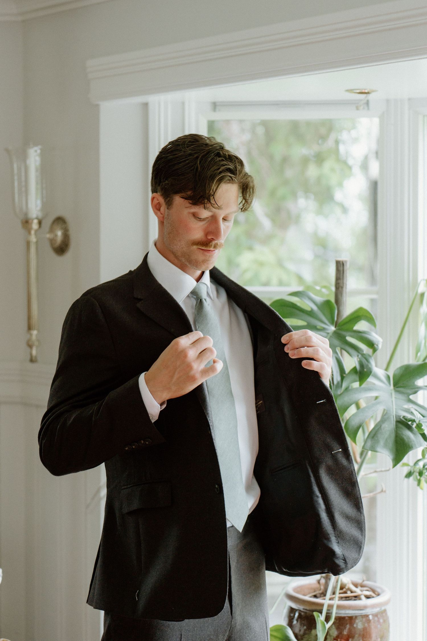 Groom in dark suit adjusting jacket lapel while standing near bright window with plants.