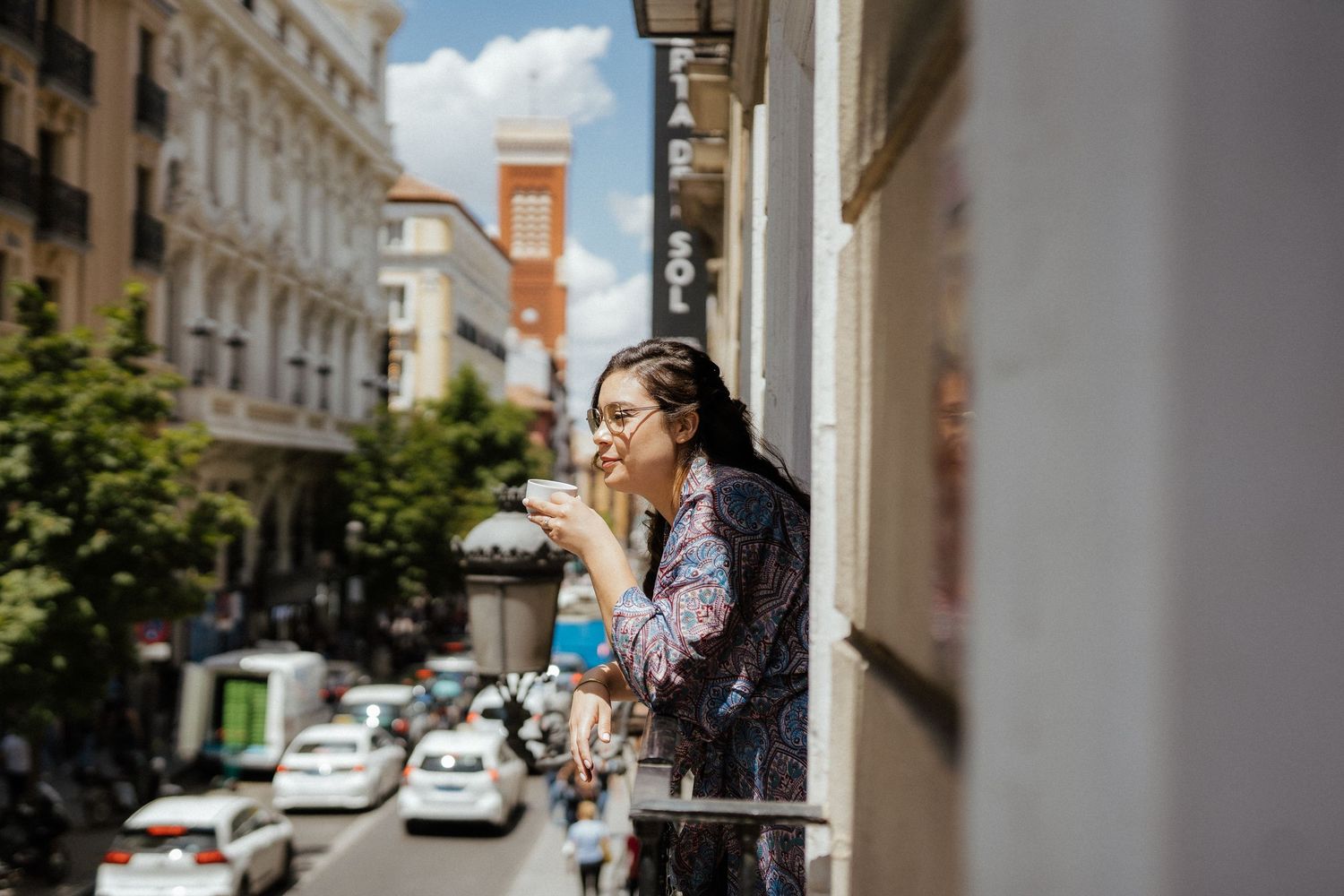 Elegant woman sips coffee on city balcony overlooking busy Madrid street on sunny day.