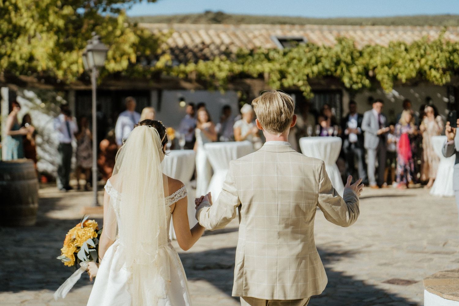 Bride and groom walk hand in hand toward outdoor wedding reception guests.