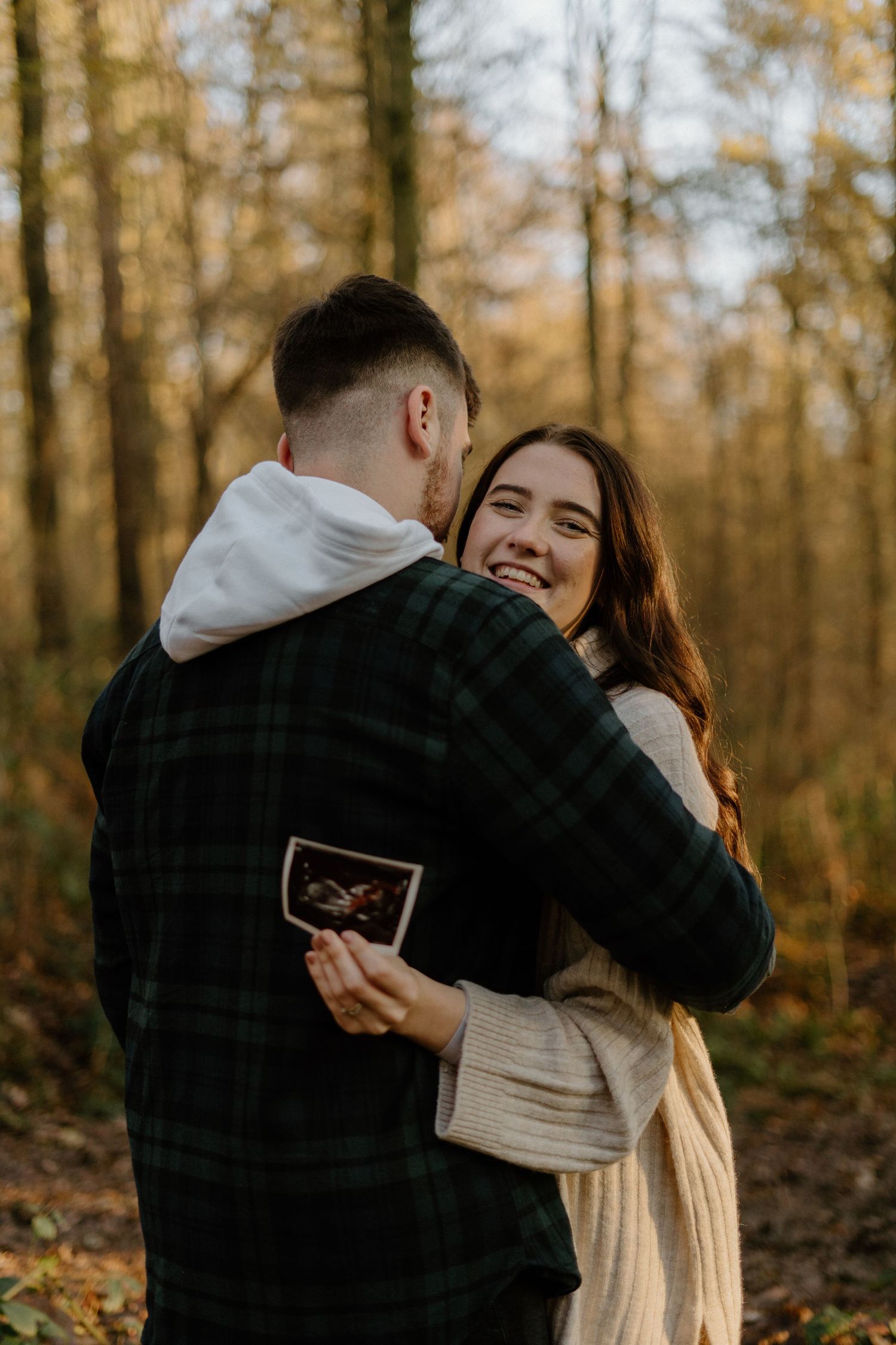 Couple hugging in autumn forest, woman smiling while holding ultrasound photo, pregnancy announcement.