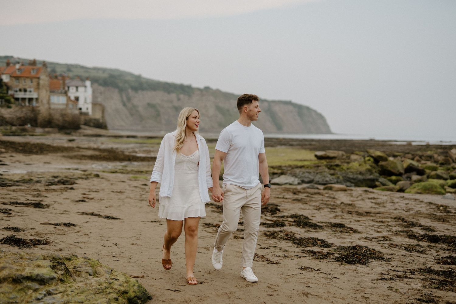 Couple walking hand in hand along a rocky beach with coastal cliffs and village behind them.