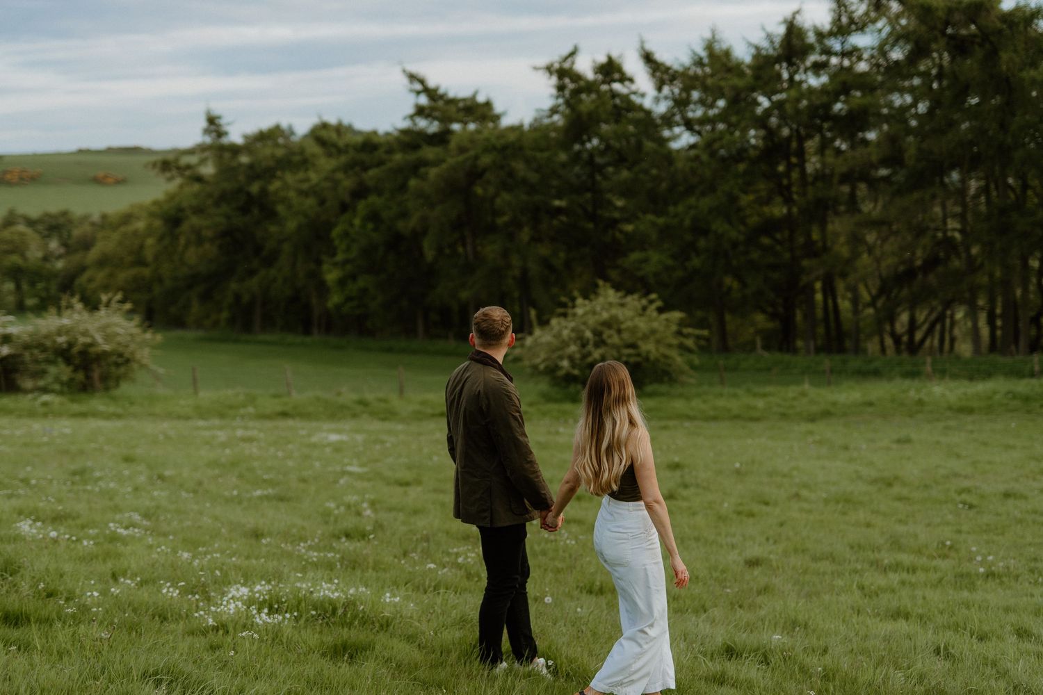 Couple holding hands walking through a lush green field with trees in the background.