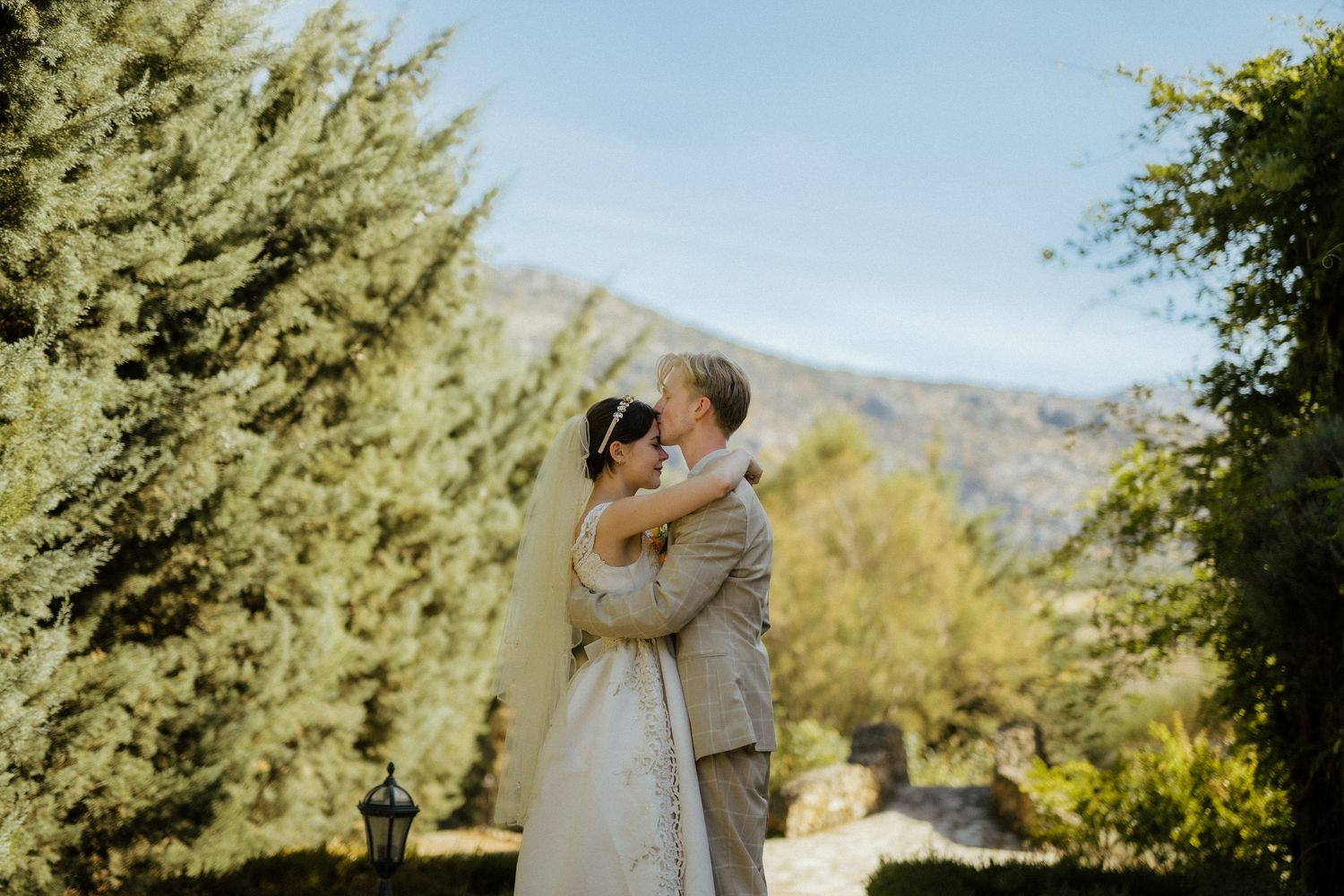 Newlyweds share romantic kiss surrounded by lush green trees and mountain scenery.