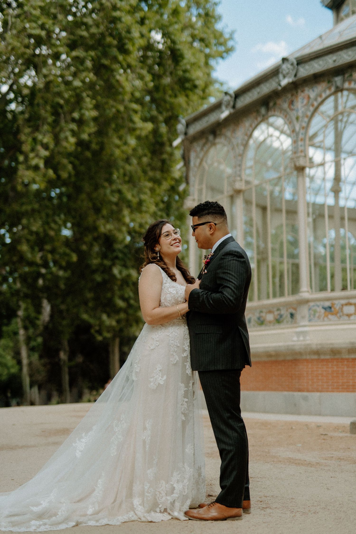 Bride and groom smiling at each other in front of a glass crystal palace surrounded by lush trees.