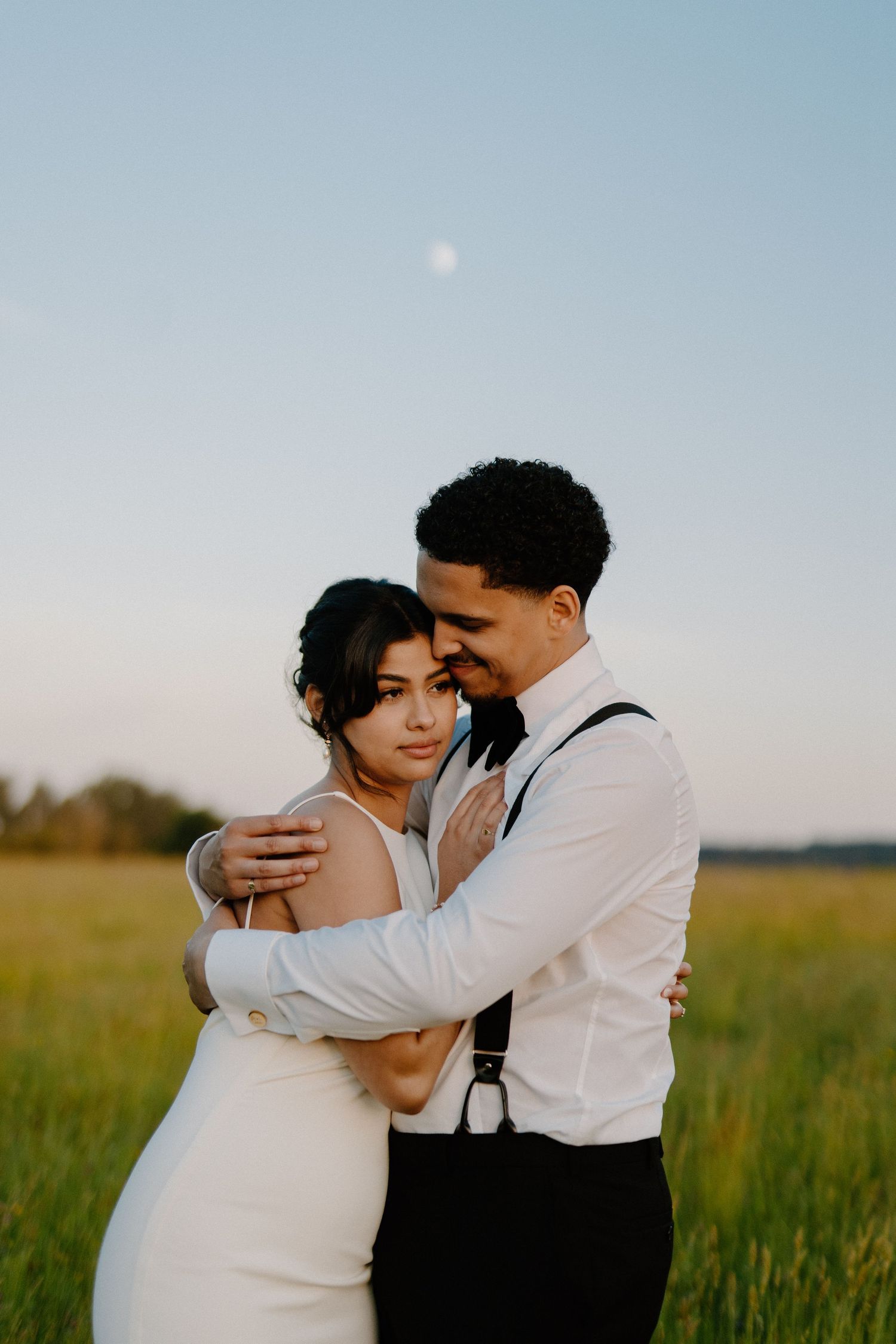 Groom tenderly embracing bride from behind in sunlit green field at golden hour.