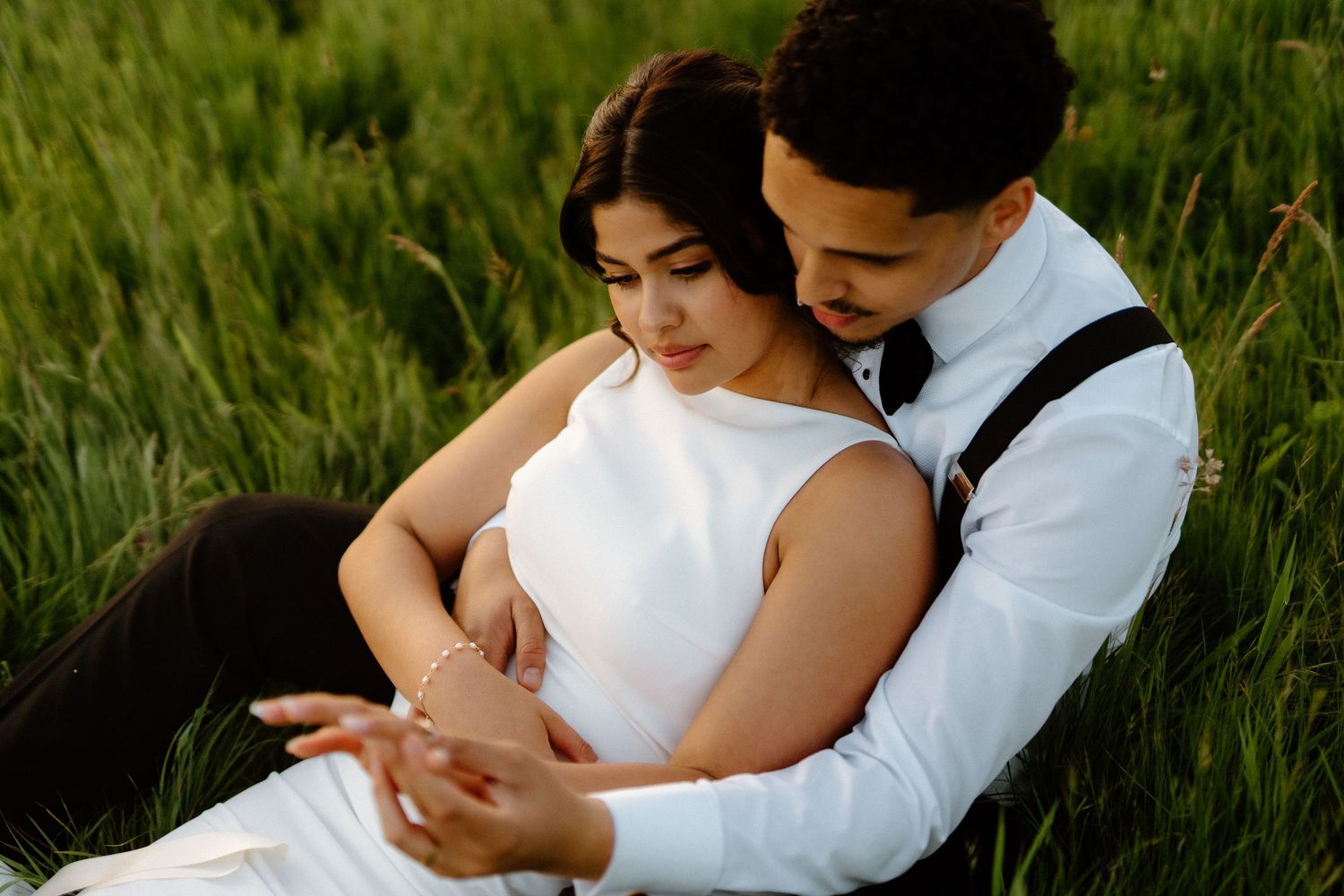 A couple in wedding attire sit closely together in a green meadow, sharing a tender romantic moment.