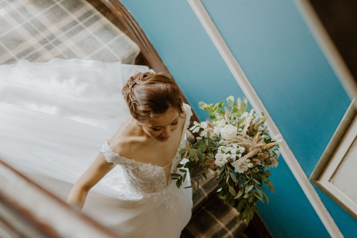 Overhead view of bride in off-shoulder lace gown carrying white and green bouquet on stairs.