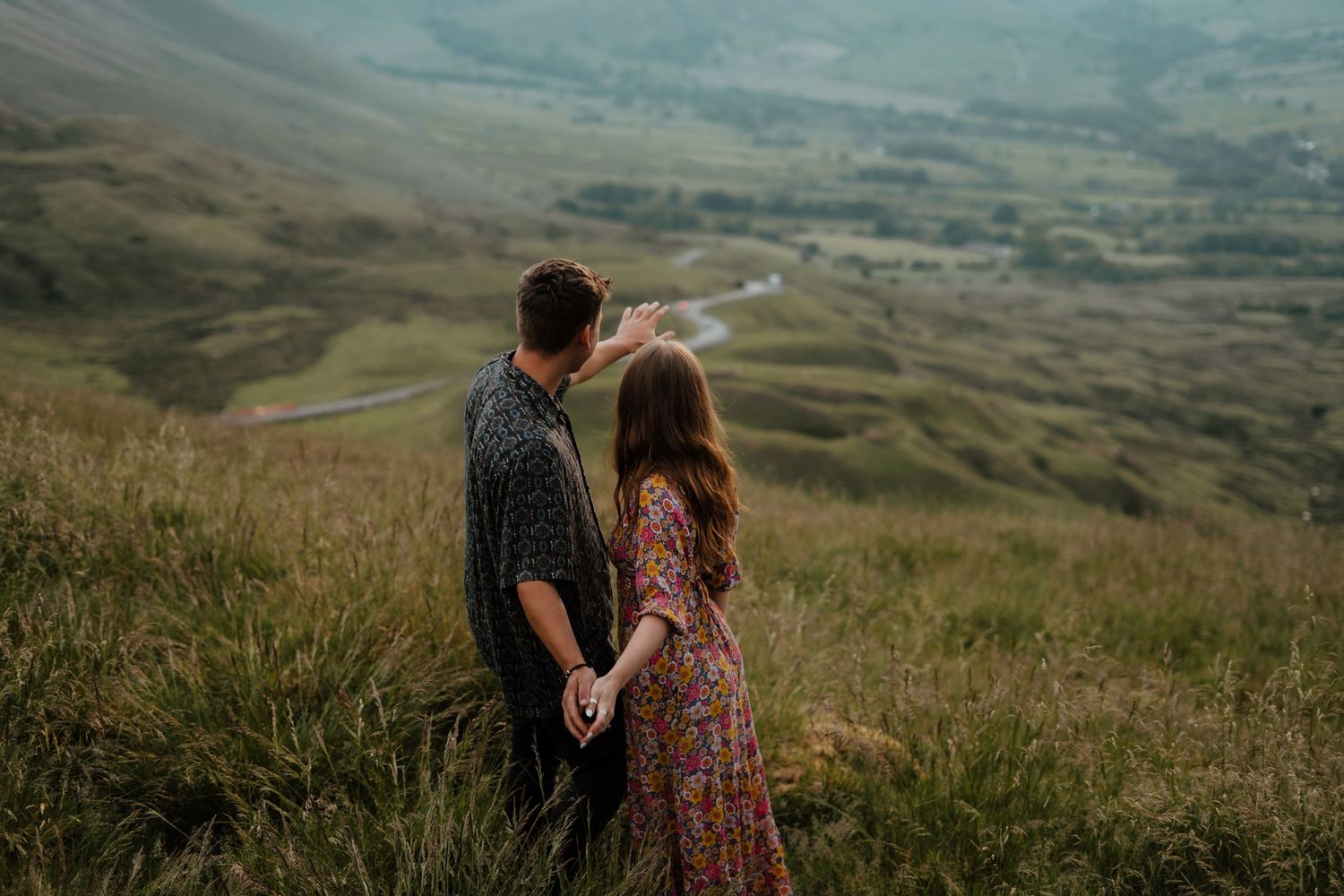 Couple holding hands while gazing at a stunning green valley landscape during engagement photos.