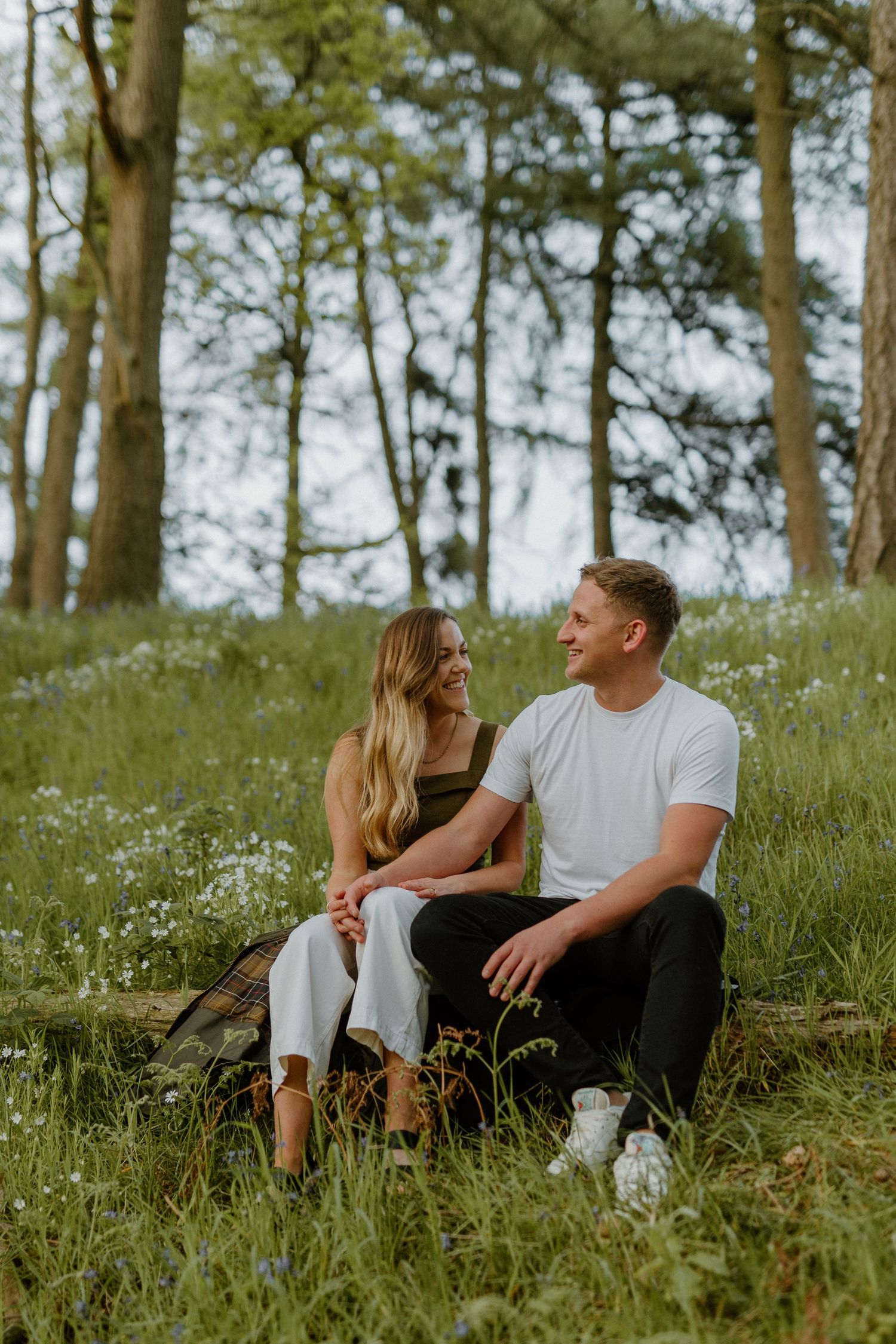 Couple sitting together in a lush green meadow with wildflowers and tall trees behind.