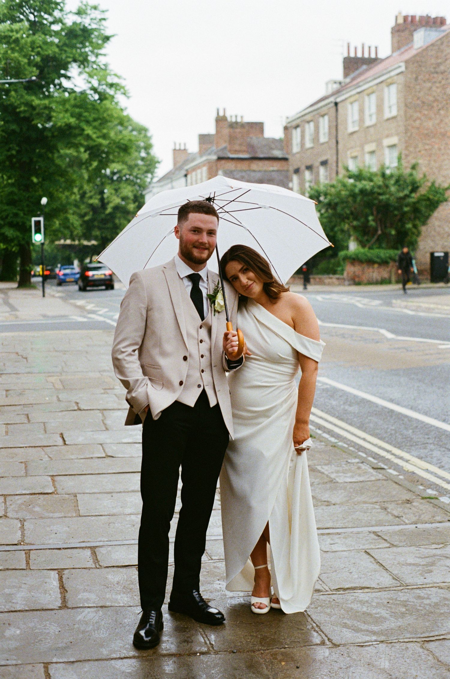 Smiling bride and groom sharing white umbrella on rainy cobblestone city street.