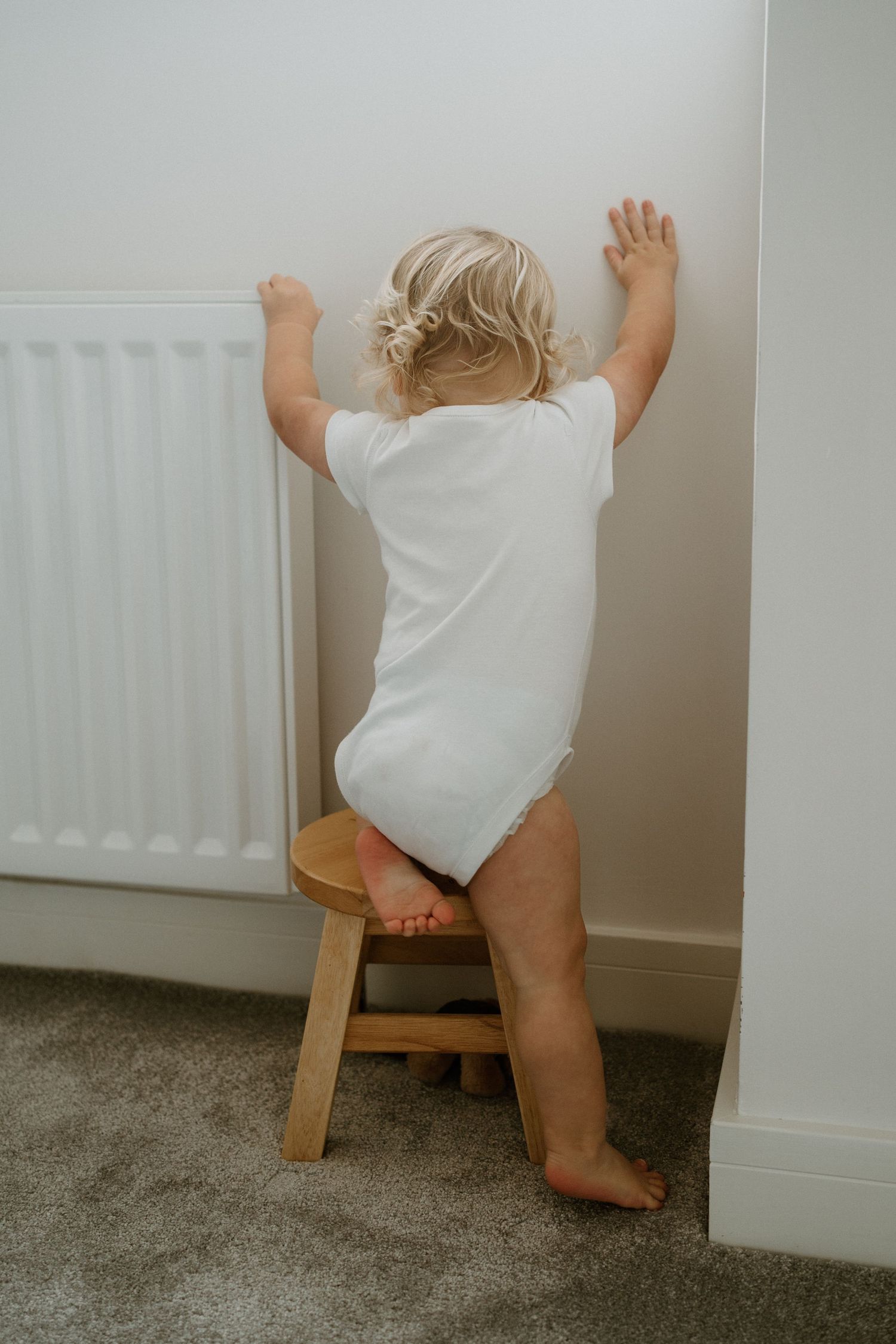 Curious toddler in white onesie standing on a wooden stool and leaning against a wall.