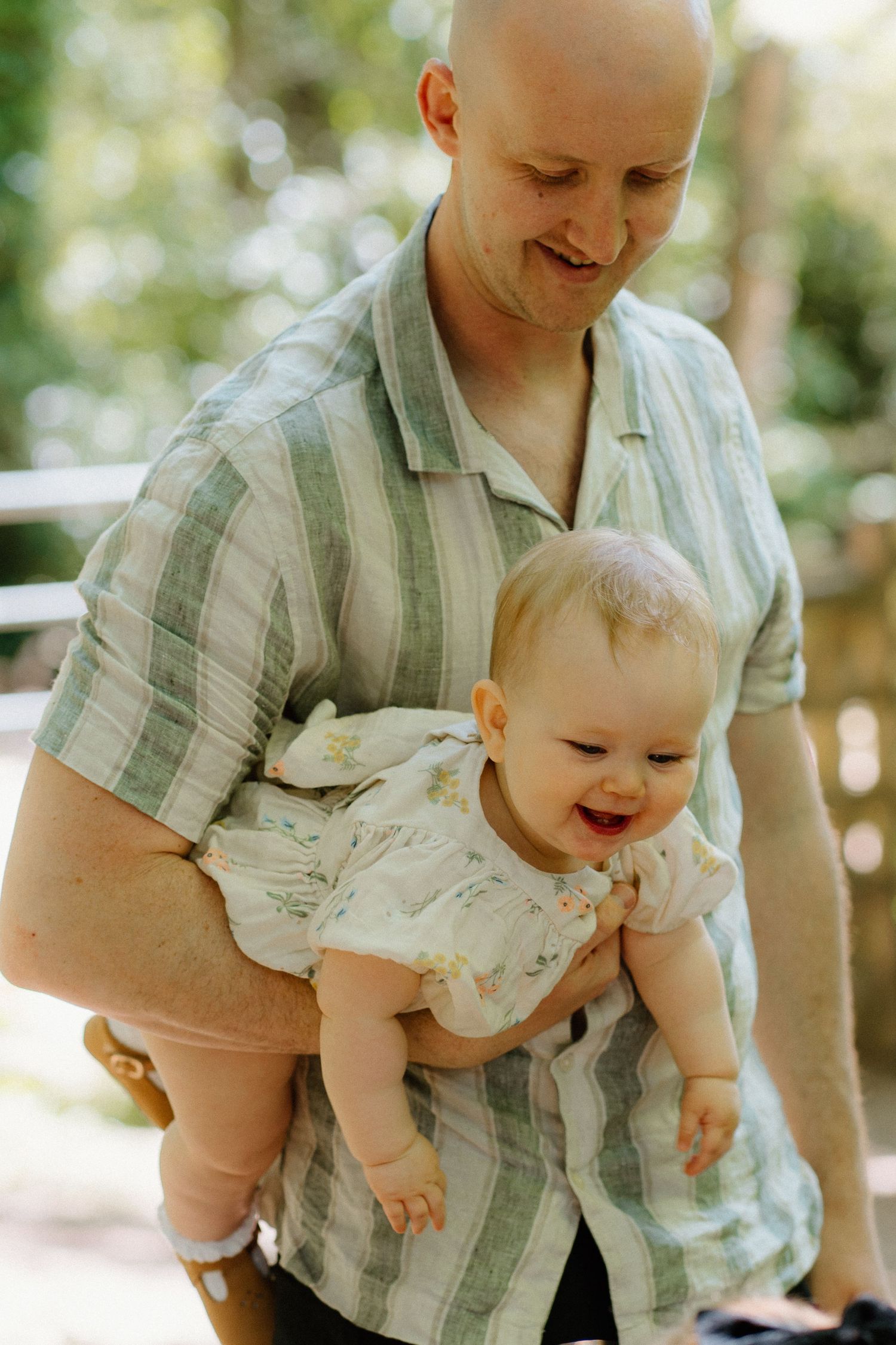 A bald father in a striped shirt holds a laughing baby outdoors in a sunny park.