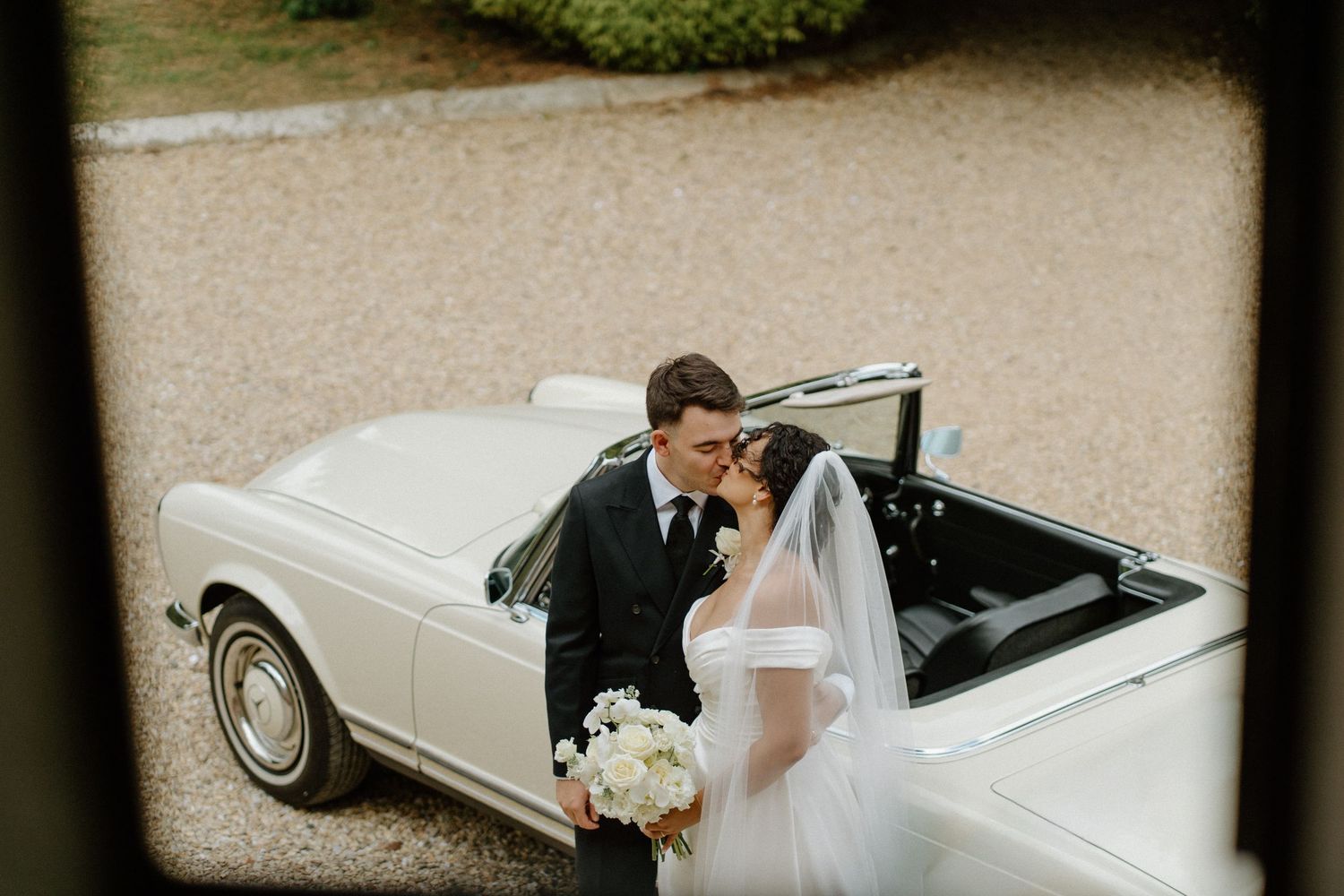 Bride and groom share tender kiss beside classic white vintage convertible car on gravel driveway.