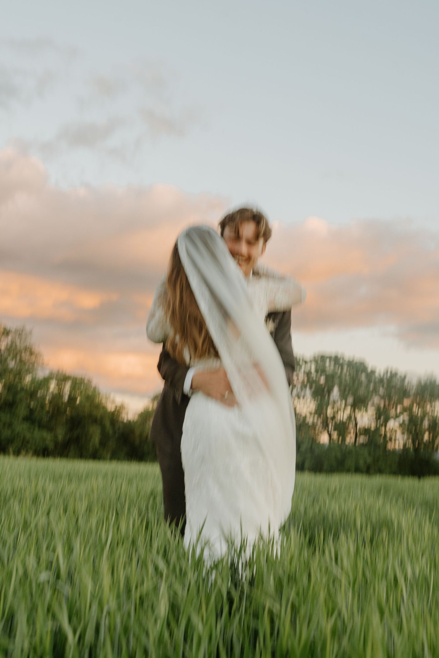 Bride and groom embrace in lush green field at golden hour sunset, veil flowing in the breeze.