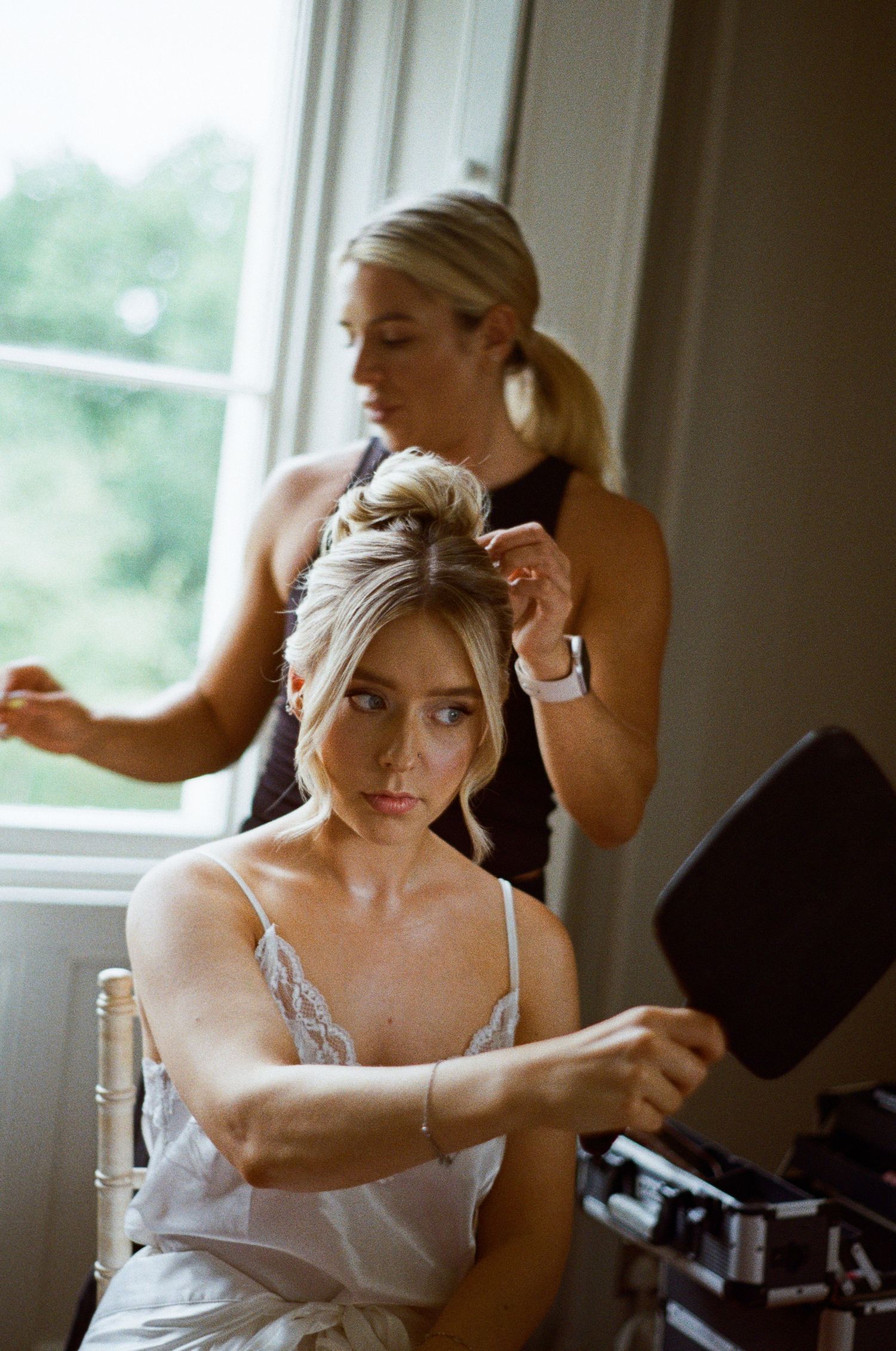 Hairstylist styling blonde bride's updo as she checks mirror during wedding morning preparations.