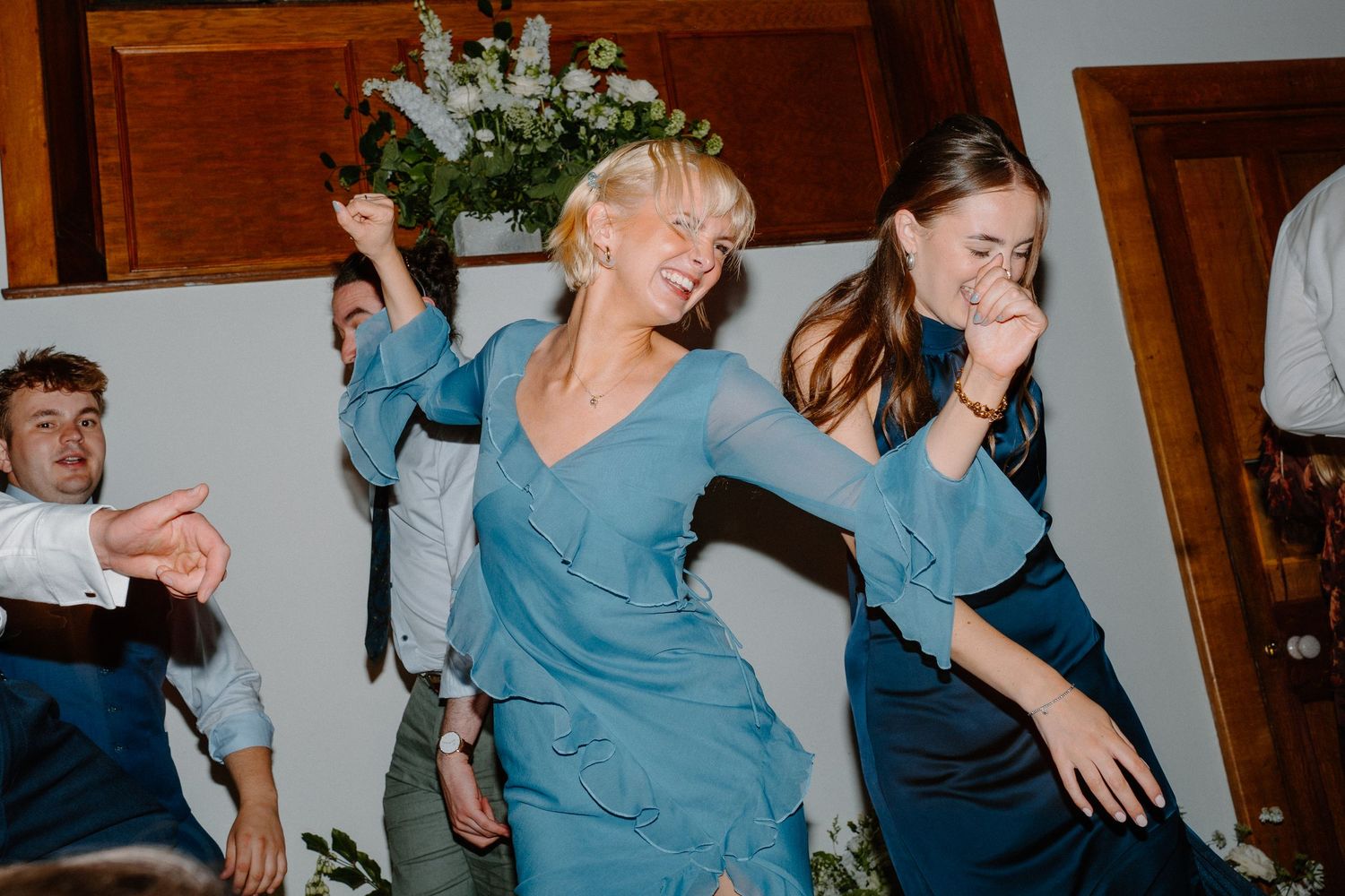 Two women in blue dresses laugh and dance joyfully together at lively wedding reception celebration.