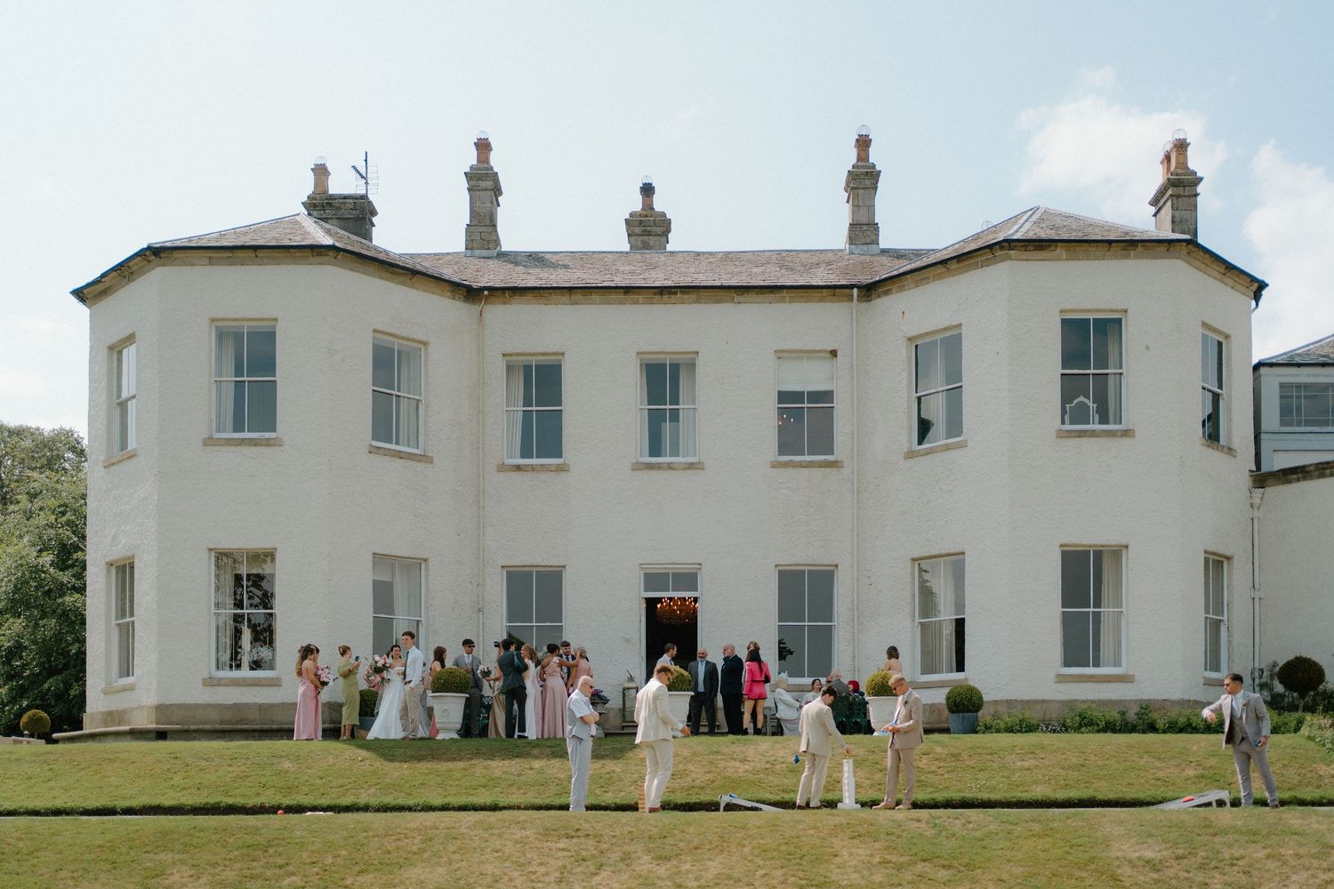 Wedding guests gathered outside a grand white Georgian country house on a sunny day.