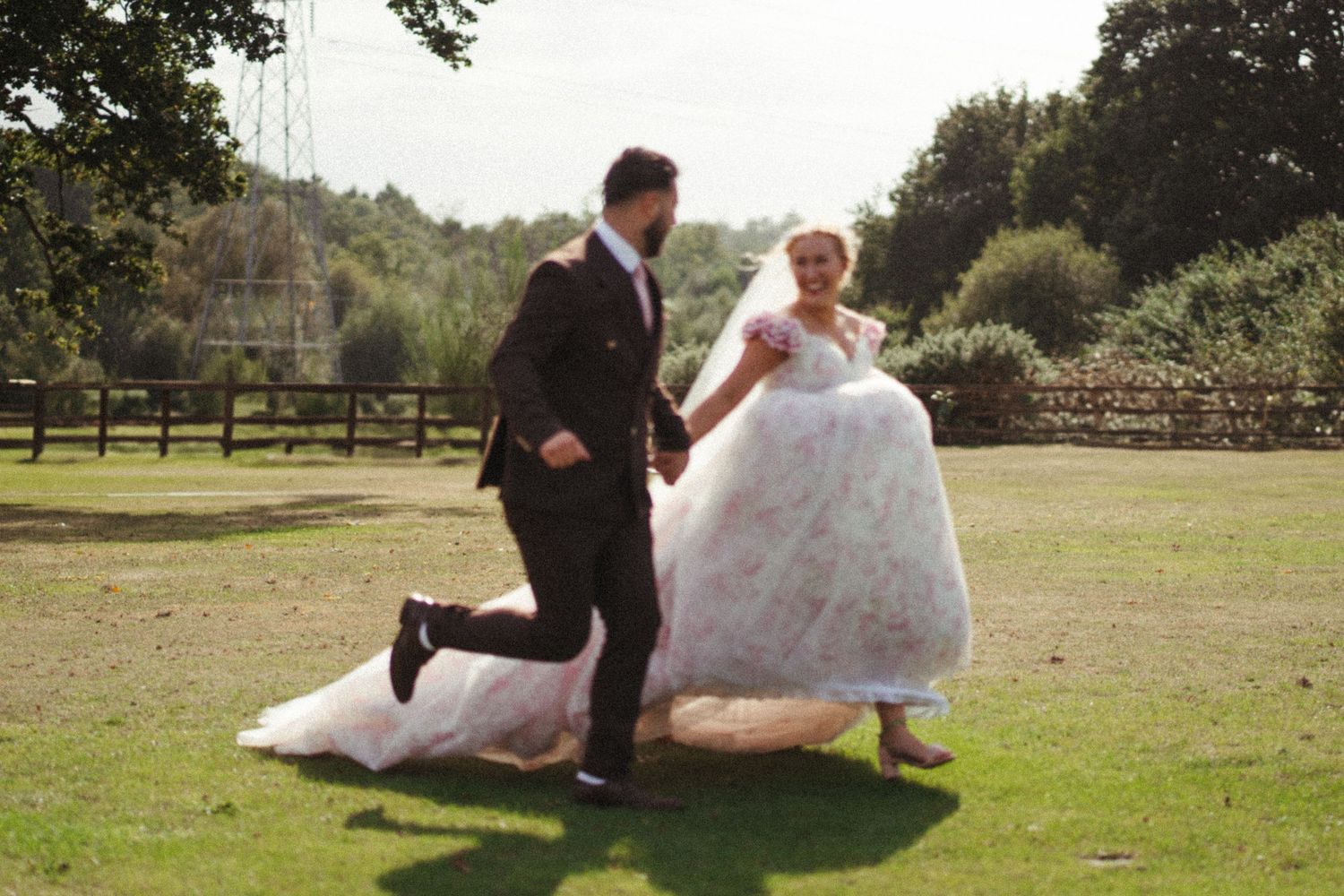 Bride in floral ball gown and groom run hand-in-hand across a sunny green lawn.