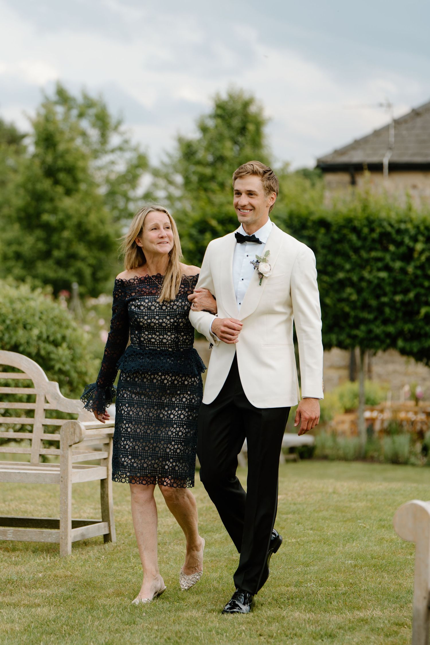Smiling groom in white tuxedo escorts elegantly dressed woman in black lace across wedding lawn.