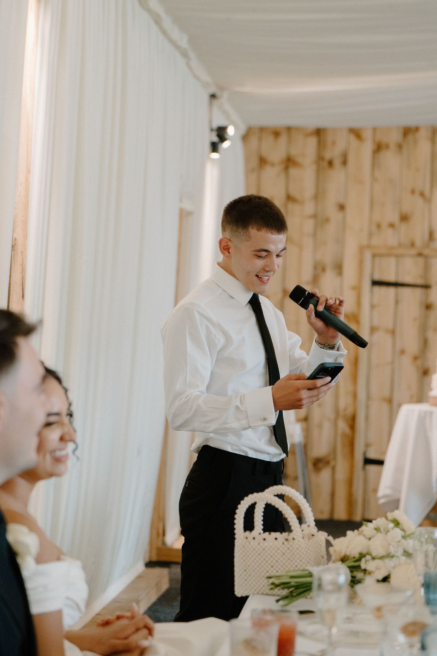 Young man in white shirt and black tie delivers a heartfelt wedding speech into a microphone.