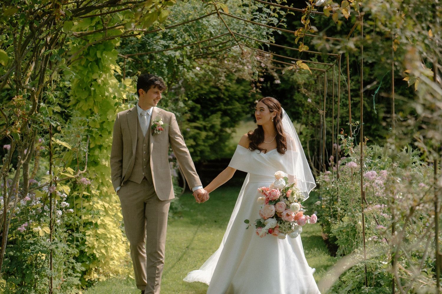 Bride in elegant white gown and groom in beige suit walking hand in hand through lush garden archway.