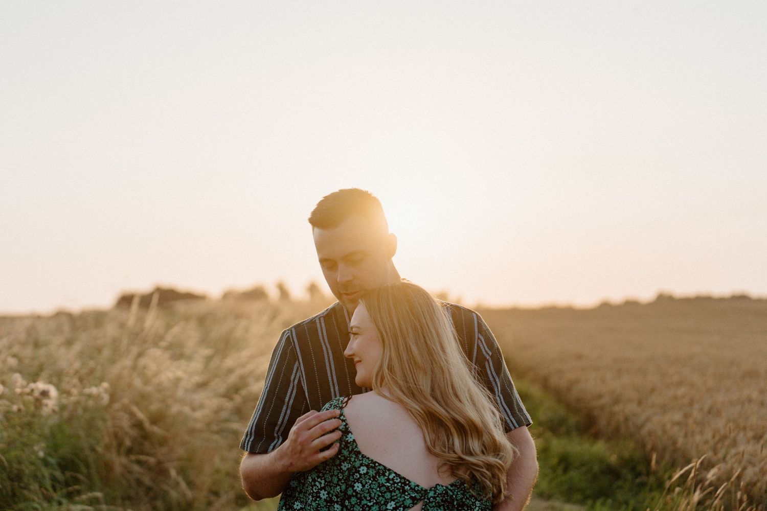 A couple embraces in a sun-drenched field at golden hour with backlit silhouettes against a warm sky.