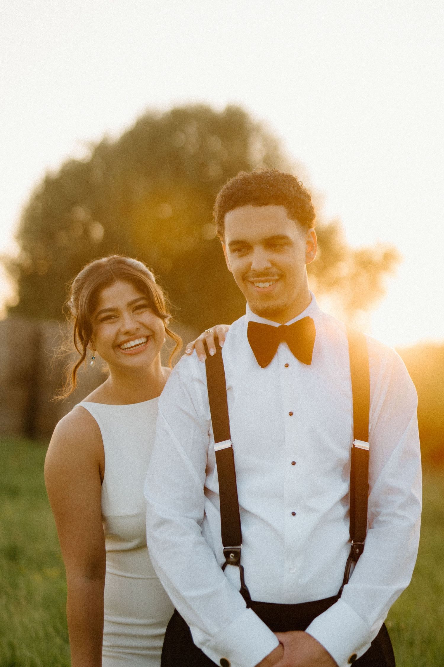 Smiling bride and groom in suspenders posing together in golden sunset light outdoors.