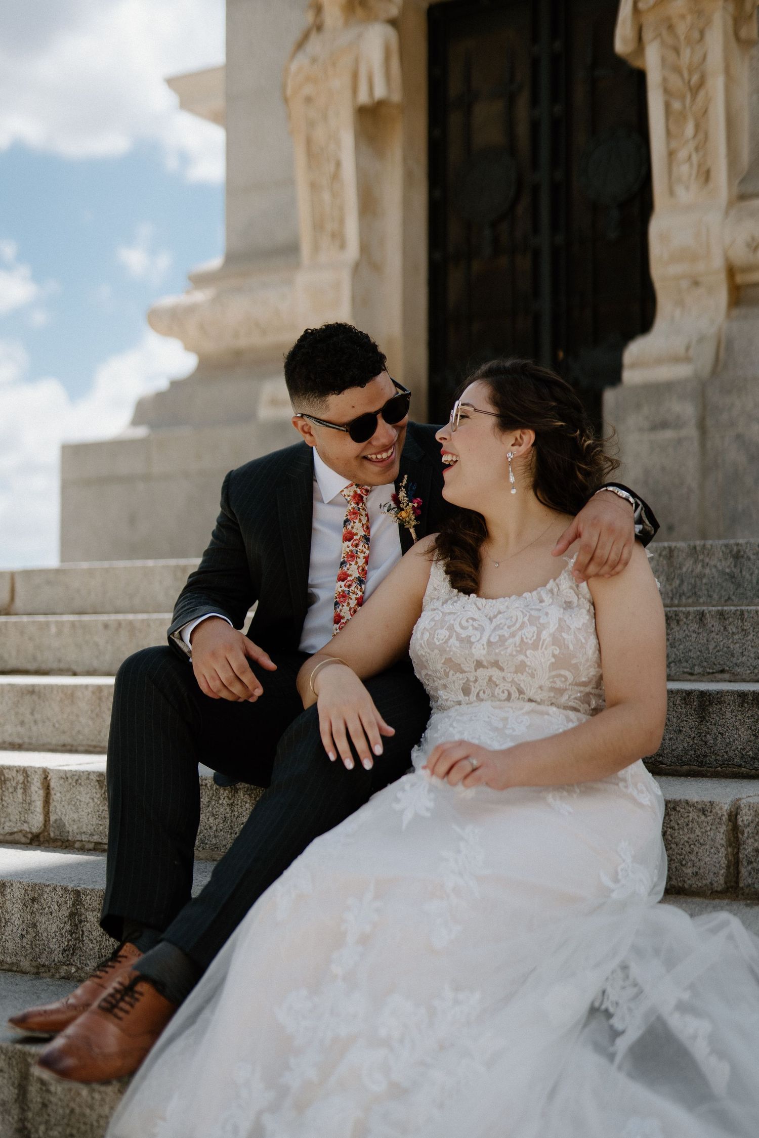Laughing bride and groom in sunglasses sitting together on stone steps of a historic monument.