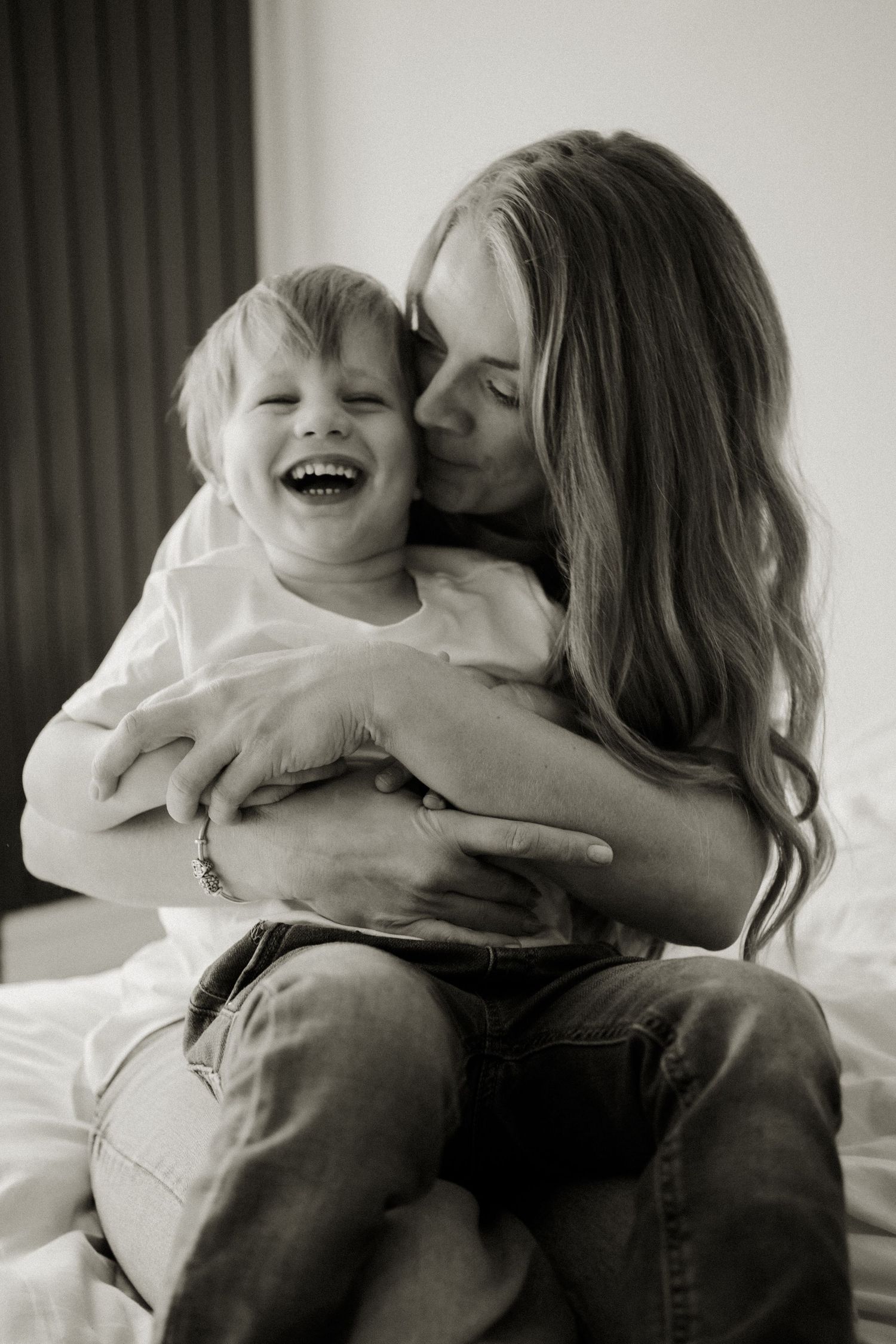 Black and white photo of a mother kissing her laughing toddler son on a bed.