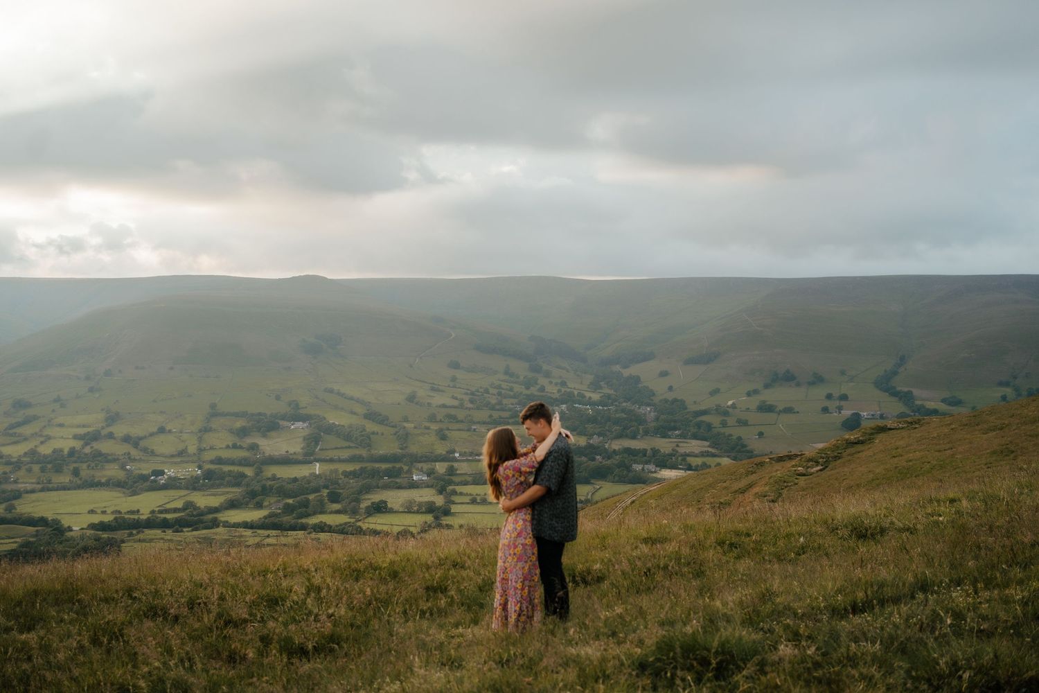 Couple sharing a tender embrace on a vast green hillside under a dramatic cloudy sky.