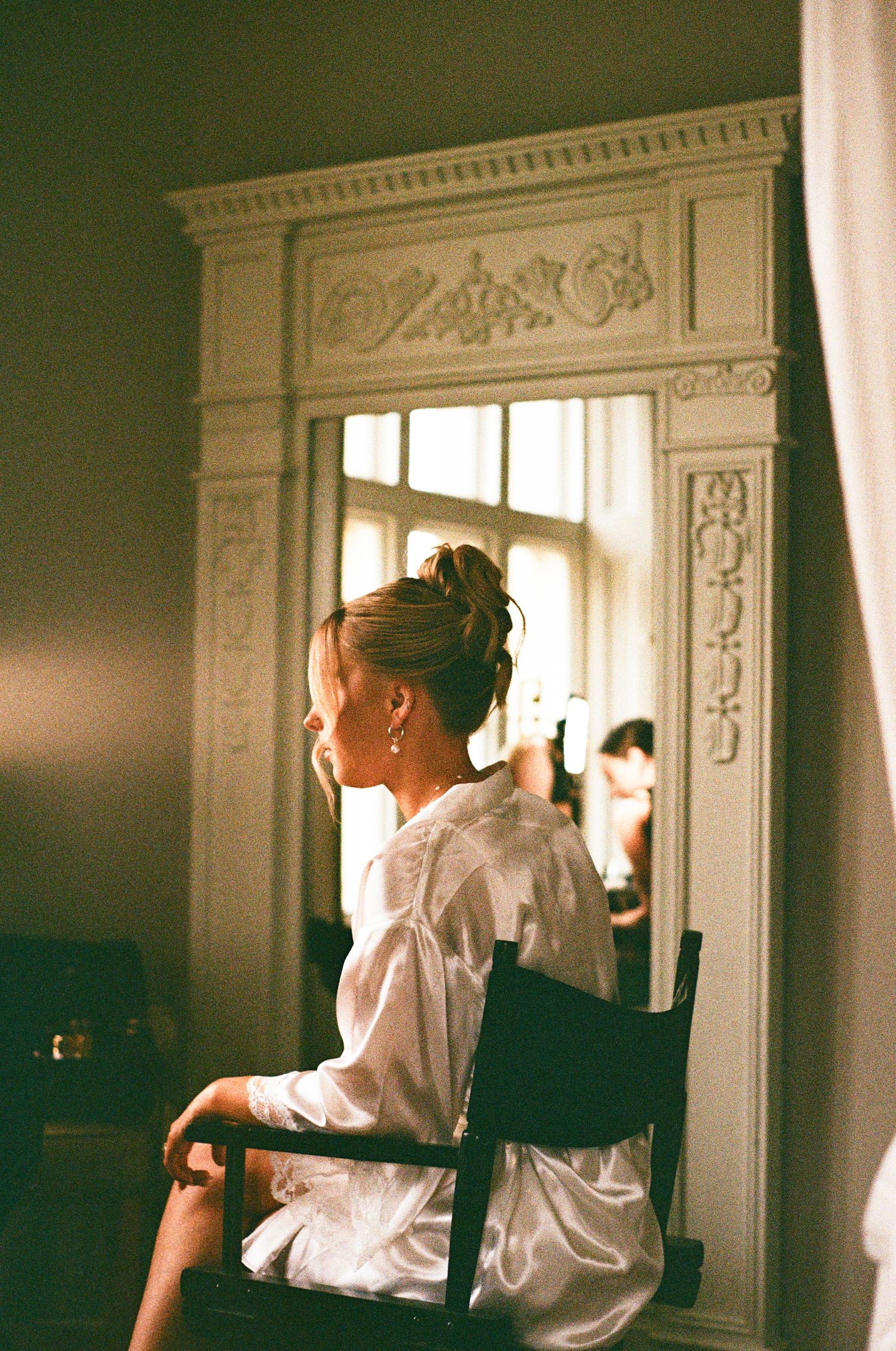 Bride in a white robe sits before an ornate mirror during wedding preparations in a vintage room.