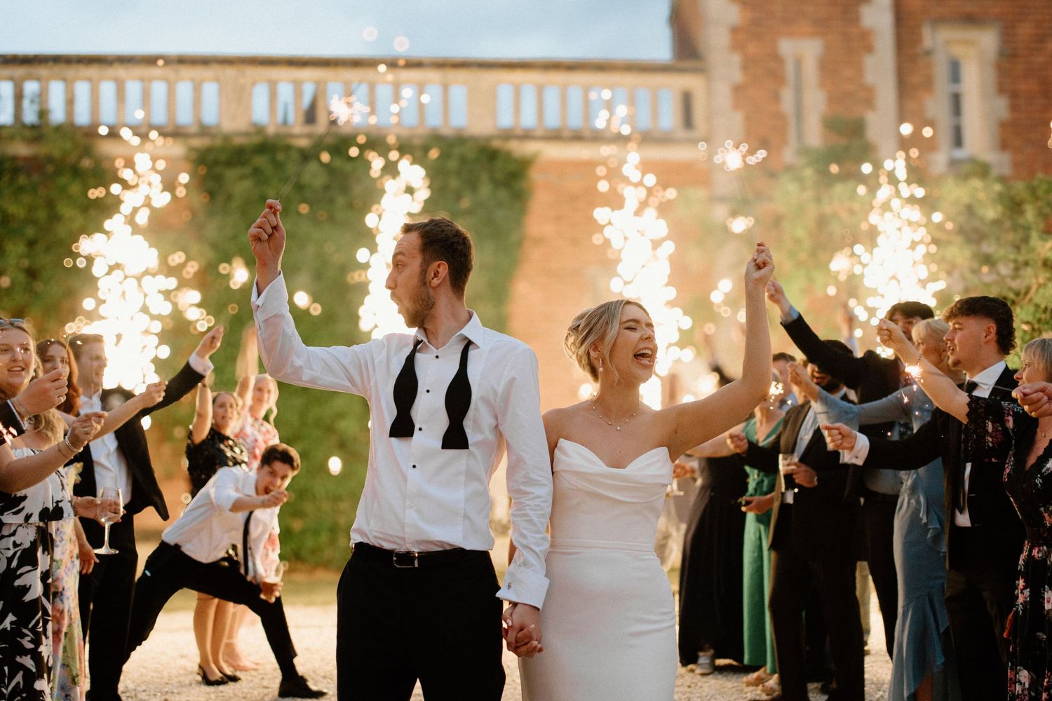 Joyful bride and groom walk through sparkler exit tunnel surrounded by cheering guests at wedding celebration.