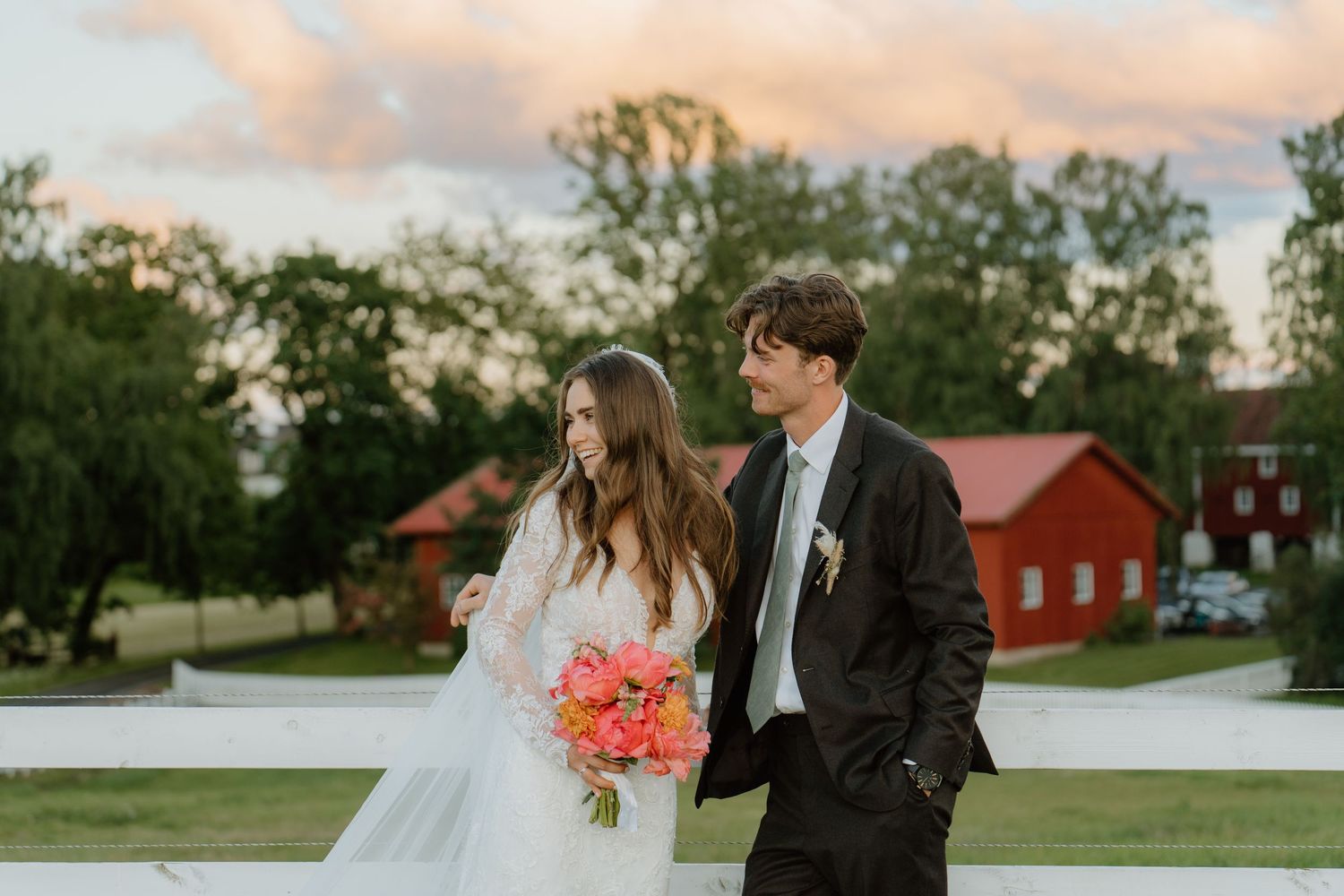 Bride and groom walk together at sunset, bride holding vibrant coral bouquet with red barn in background.