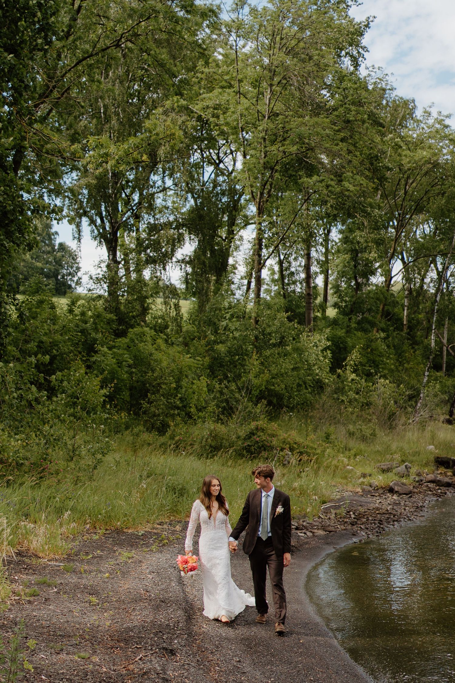 Bride and groom walking hand in hand on scenic path beside river and green trees.