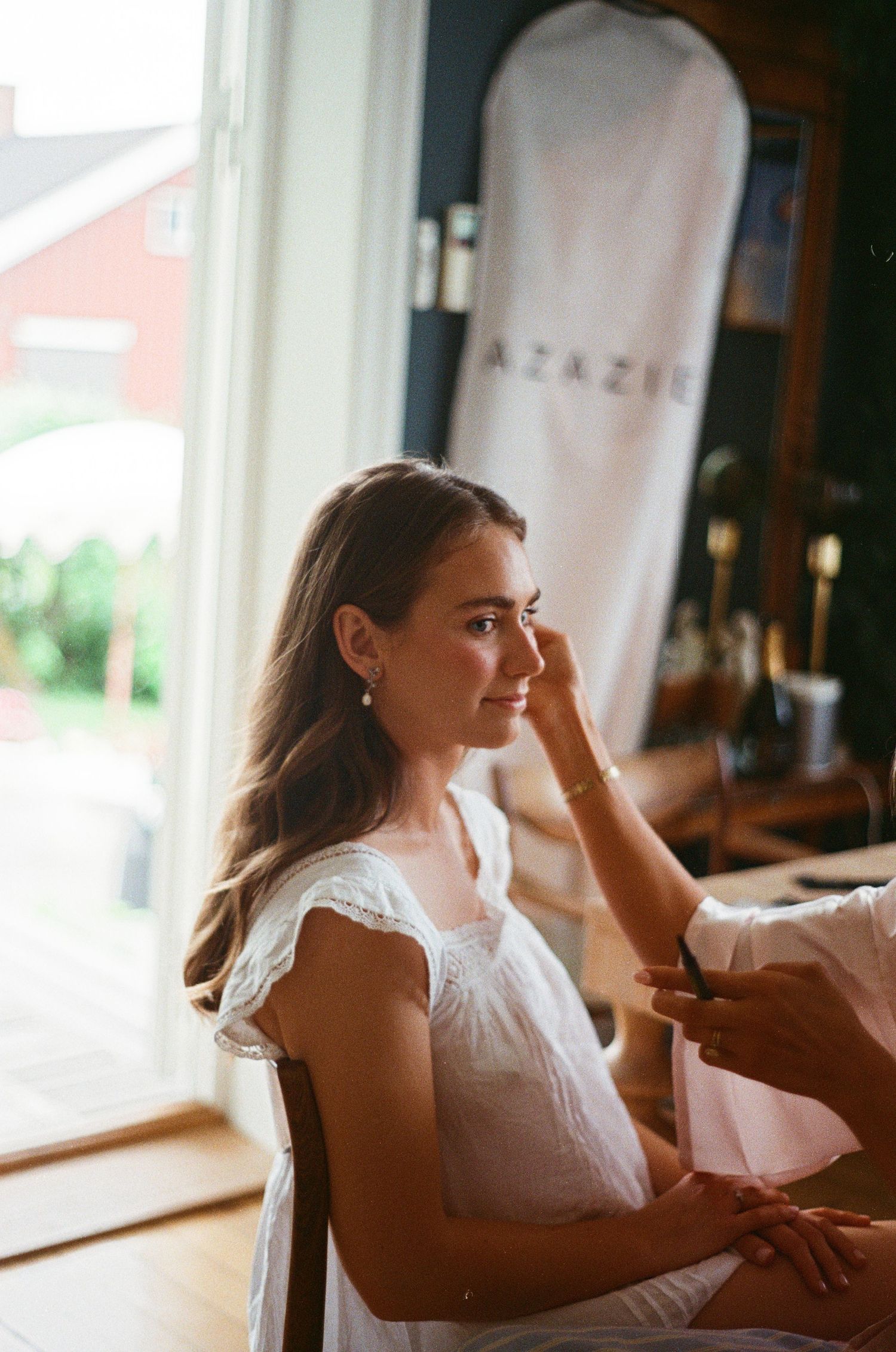 Bride getting makeup applied while seated near a bright window on her wedding morning.